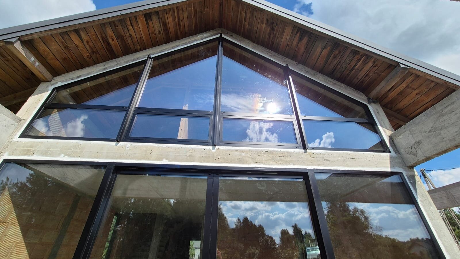 Duże okna z czarnymi ramami w surowym stanie budynku, odbijające niebo i drzewa. Trapezowe okno na szczycie. Widoczne drewniane elementy dachu i beton konstrukcyjny.