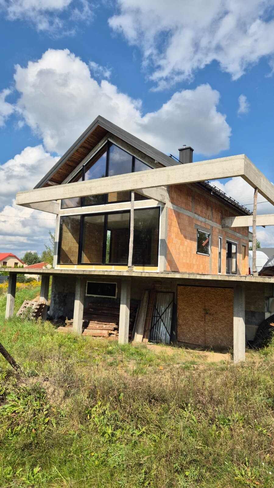 Surowy stan domu z cegły z dużymi oknami i betonowym zadaszeniem, w tle błękitne niebo z chmurami. Widoczne elementy konstrukcyjne i wykończeniowe.