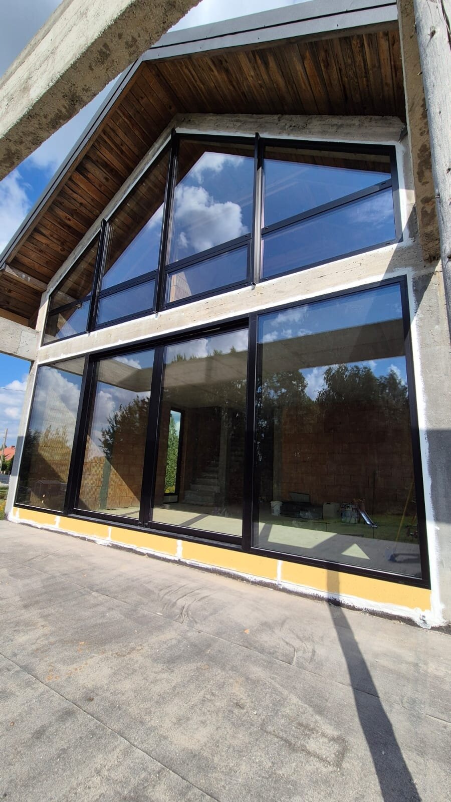 Nowoczesna fasada budynku w stanie surowym z dużymi, czarnymi oknami. Widoczne odbicie nieba i chmur w szybach. Surowy beton i drewniane elementy konstrukcyjne.