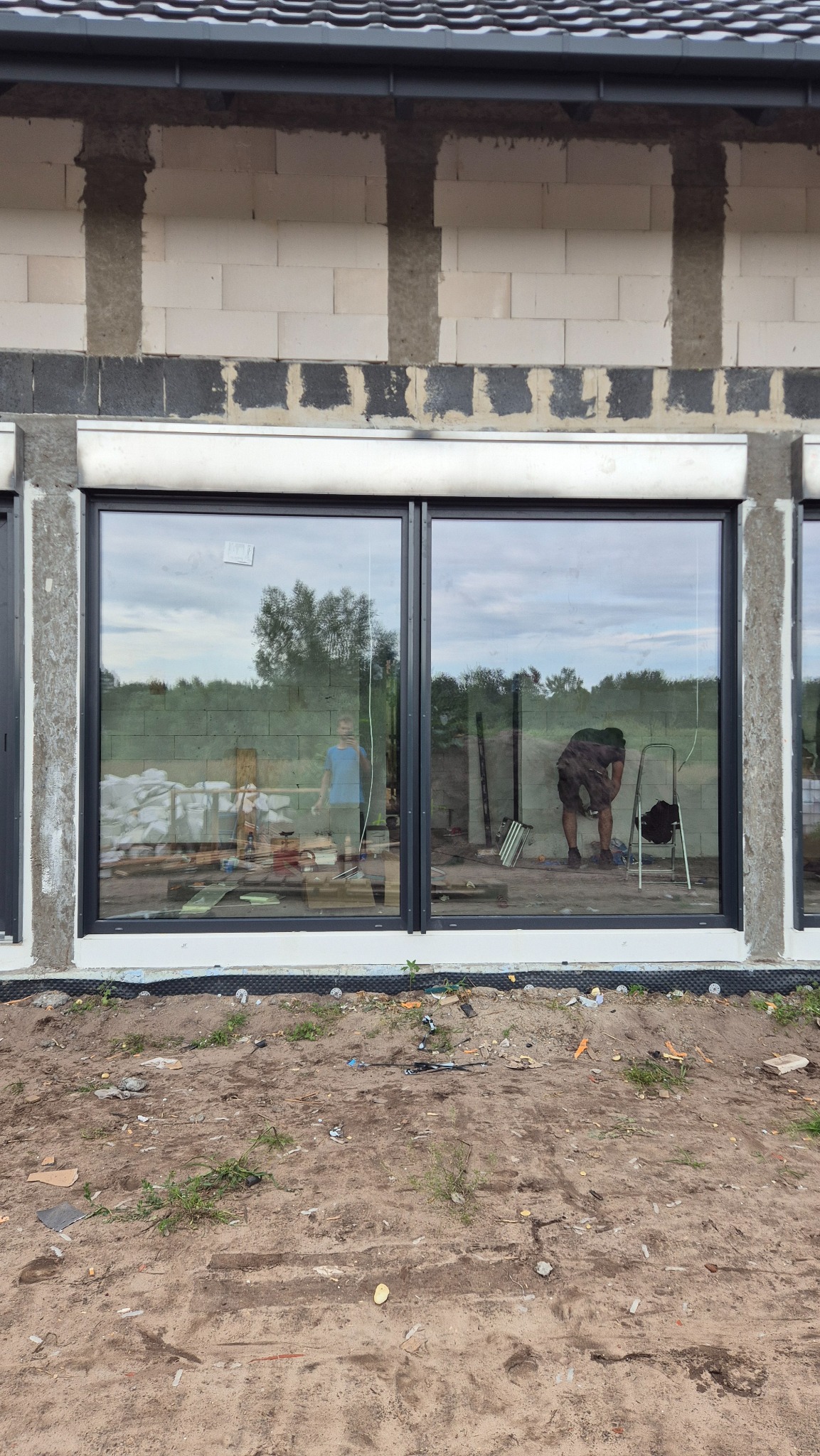 Montaż okien z czarnymi ramami w surowym budynku. Widoczne narzędzia i pracownicy w odbiciu szyb. Okna z roletami zewnętrznymi.