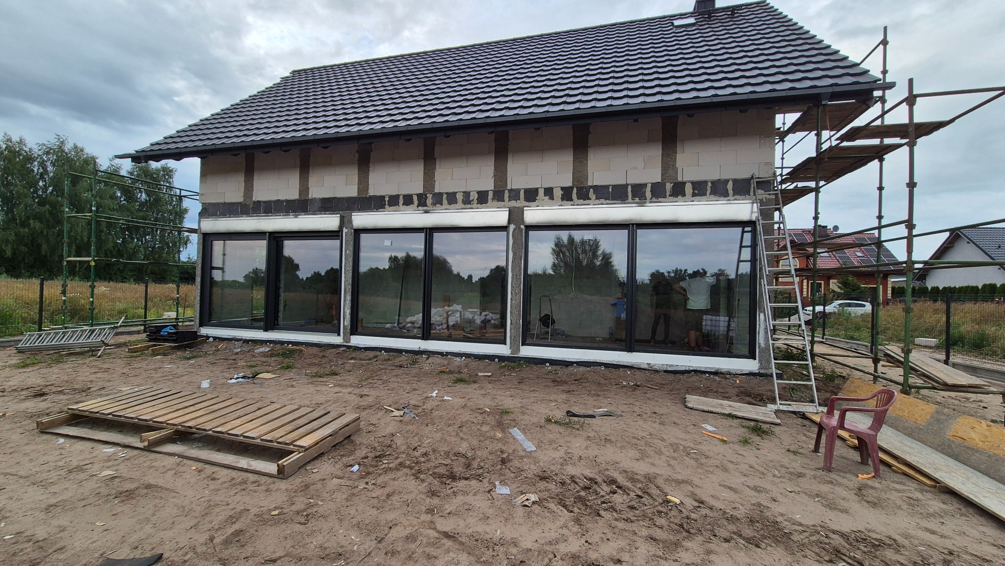 Surowy stan budynku z dużymi oknami w czarnych ramach, widoczne rusztowania i elementy budowlane na zewnątrz. W oknach odbija się krajobraz.