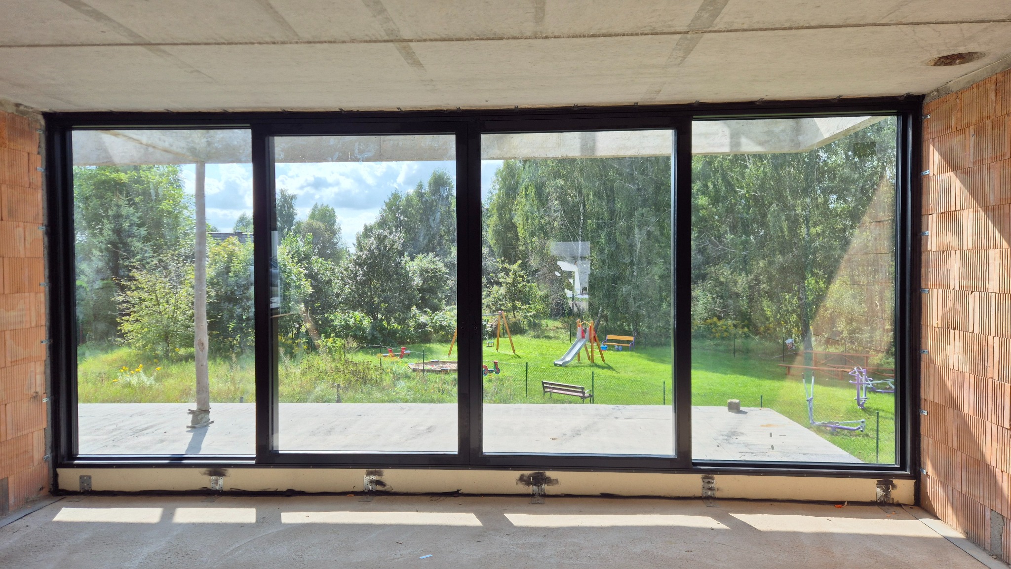 Duże, czarne okno tarasowe z widokiem na plac zabaw i zieleń, zamontowane w surowym wnętrzu z ceglanymi ścianami i betonową podłogą. Widoczne elementy montażowe.