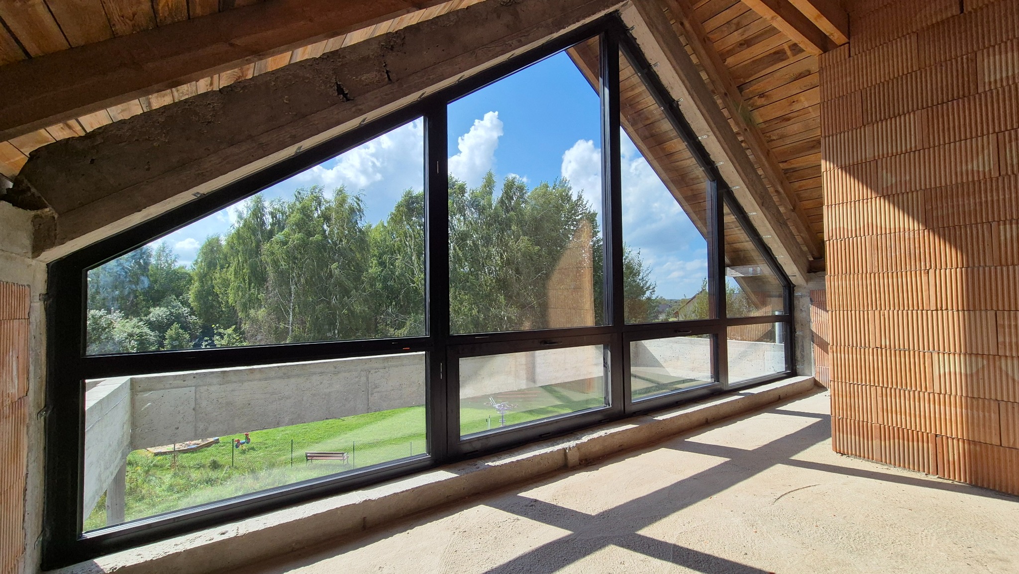 Duże okno z czarną ramą w surowym wnętrzu poddasza, widok na zieleń i błękitne niebo. Cień rzucany na podłogę. Cegła dziurawka na ścianie.