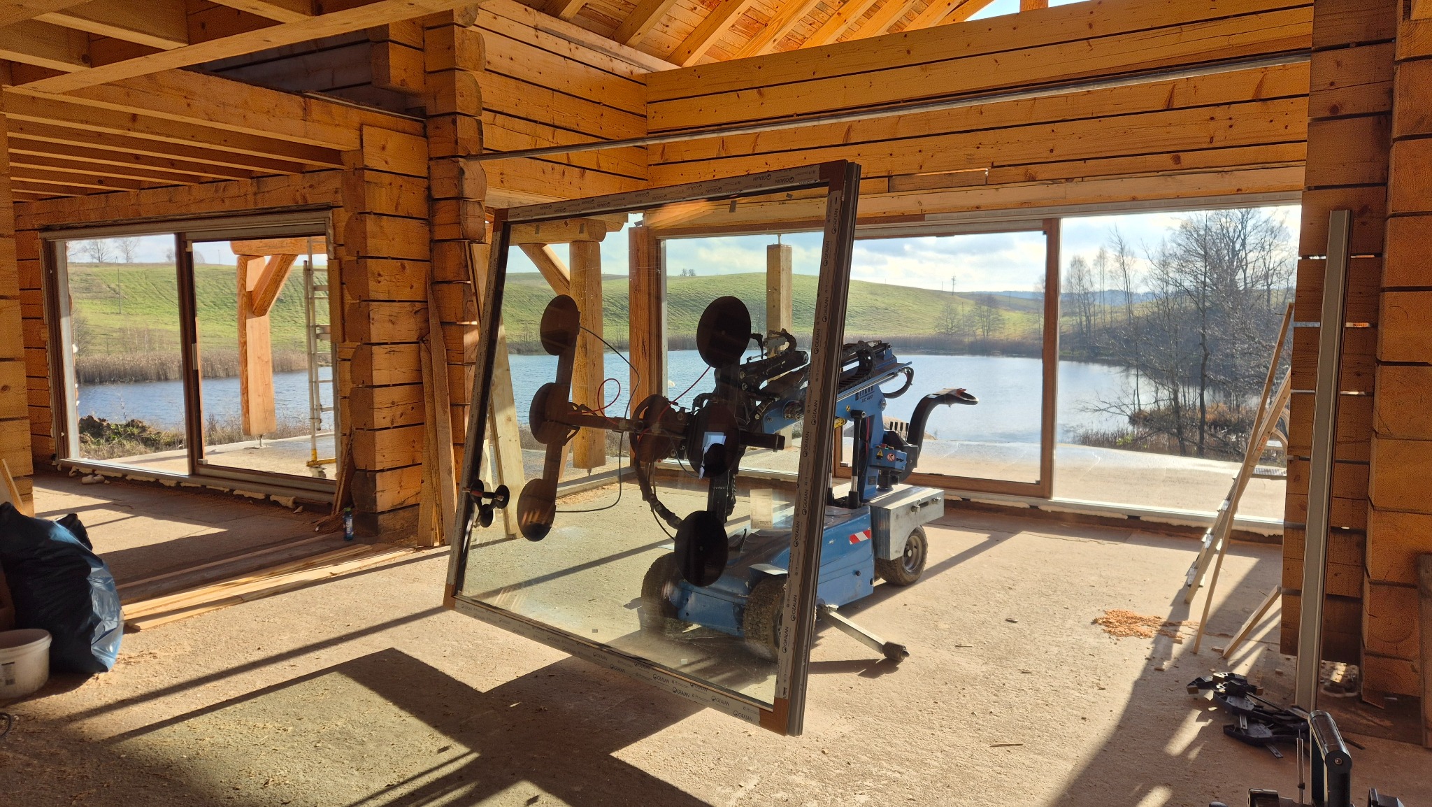 Montaż okna w drewnianym domu z widokiem na jezioro. Specjalistyczny sprzęt do przenoszenia szyb. Jasne wnętrze w surowym stanie.