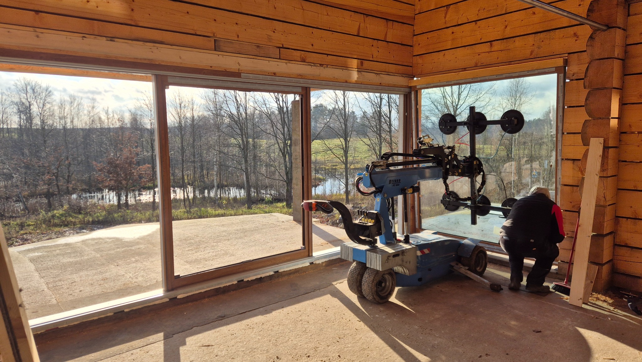 Montaż panoramicznego okna w drewnianym domu z widokiem na krajobraz. Użycie specjalistycznego robota do przenoszenia szyb. Praca w toku, widoczny pracownik.