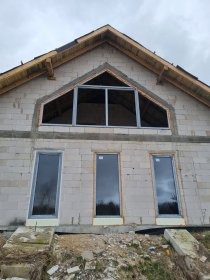 Surowy stan budynku z zamontowanymi oknami PCV o grafitowej ramie. Widoczne trzy okna pionowe i jedno trójkątne na poddaszu. Budynek w trakcie budowy.