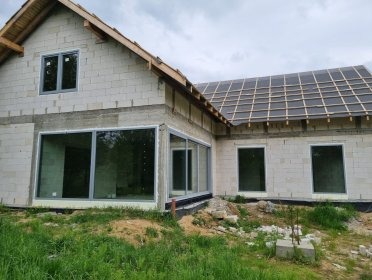 Surowy stan domu z dużymi oknami w Morągu. Widoczne szare ramy okienne i fragment dachu z czarną membraną. Budynek w trakcie budowy, otoczony zielenią.