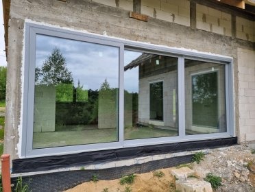 Szerokie, przesuwne okno z szarymi ramami w surowym stanie budynku. Widok na zewnątrz odbija się w szybach, ukazując zieleń i fragment domu.