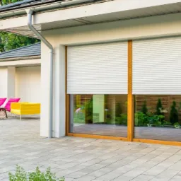 Elewacja domu z dużym oknem zabezpieczonym białą roletą zewnętrzną, widoczny fragment ogrodu z domkiem na drzewie i kolorowymi leżakami.