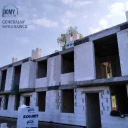 Budynek w stanie surowym otwartym z białych bloczków, widoczne otwory okienne i drzwiowe, paleta bloczków Solbet na pierwszym planie, logo H+H na górze budynku, niebieskie niebo w tle.