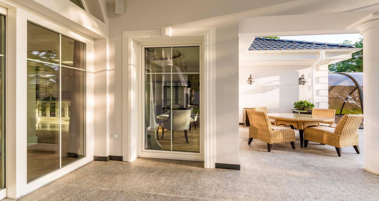 Nowoczesne, białe okna z podziałami szprosowymi w eleganckim domu z tarasem i rattanowymi meblami ogrodowymi widocznymi w tle.
