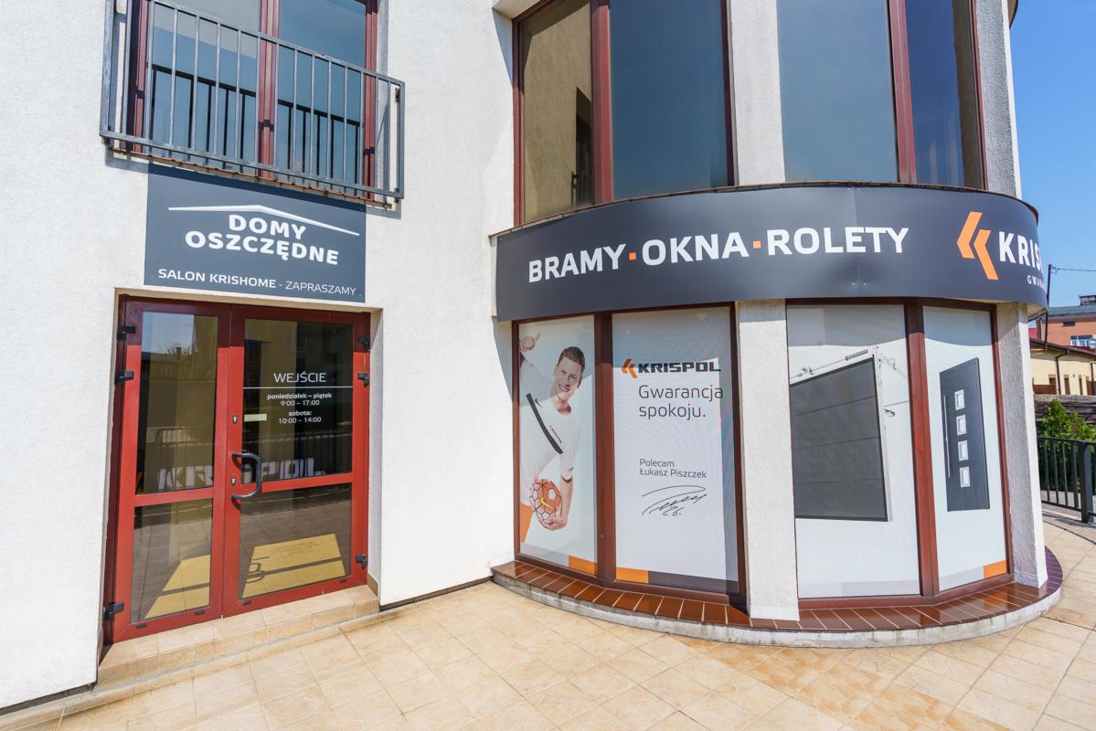 Fasada salonu sprzedaży 'Domy Oszczędne' z ekspozycją bram, okien i rolet, widoczne godziny otwarcia i reklama firmy Krispol z gwarancją spokoju.
