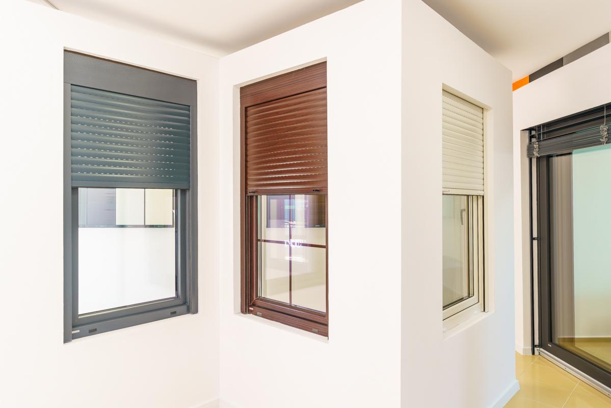 Wnętrze z trzema różnymi oknami z roletami zewnętrznymi w kolorach: szarym, brązowym i kremowym, zamontowanymi w białych ścianach.