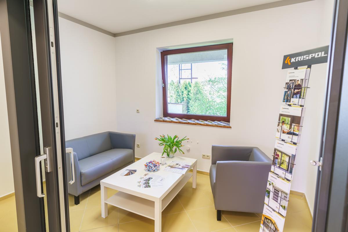 Jasne wnętrze poczekalni z szarą sofą, fotelem i stolikiem kawowym, okno z brązową ramą i ulotki reklamowe firmy Krispol.