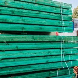 Stos zielonych, impregnowanych desek budowlanych ułożonych na składzie, zabezpieczonych białym sznurkiem, w tle widoczne inne materiały budowlane i budynek magazynowy.