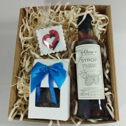 Endi-Mar Sp. z o.o. - Kartonowy kosz prezentowy wypełniony ozdobnym siankiem, zawierający syrop malinowo-imbirowy, herbatę w białym pudełku z niebieską kokardą i piernik w kształcie serca.