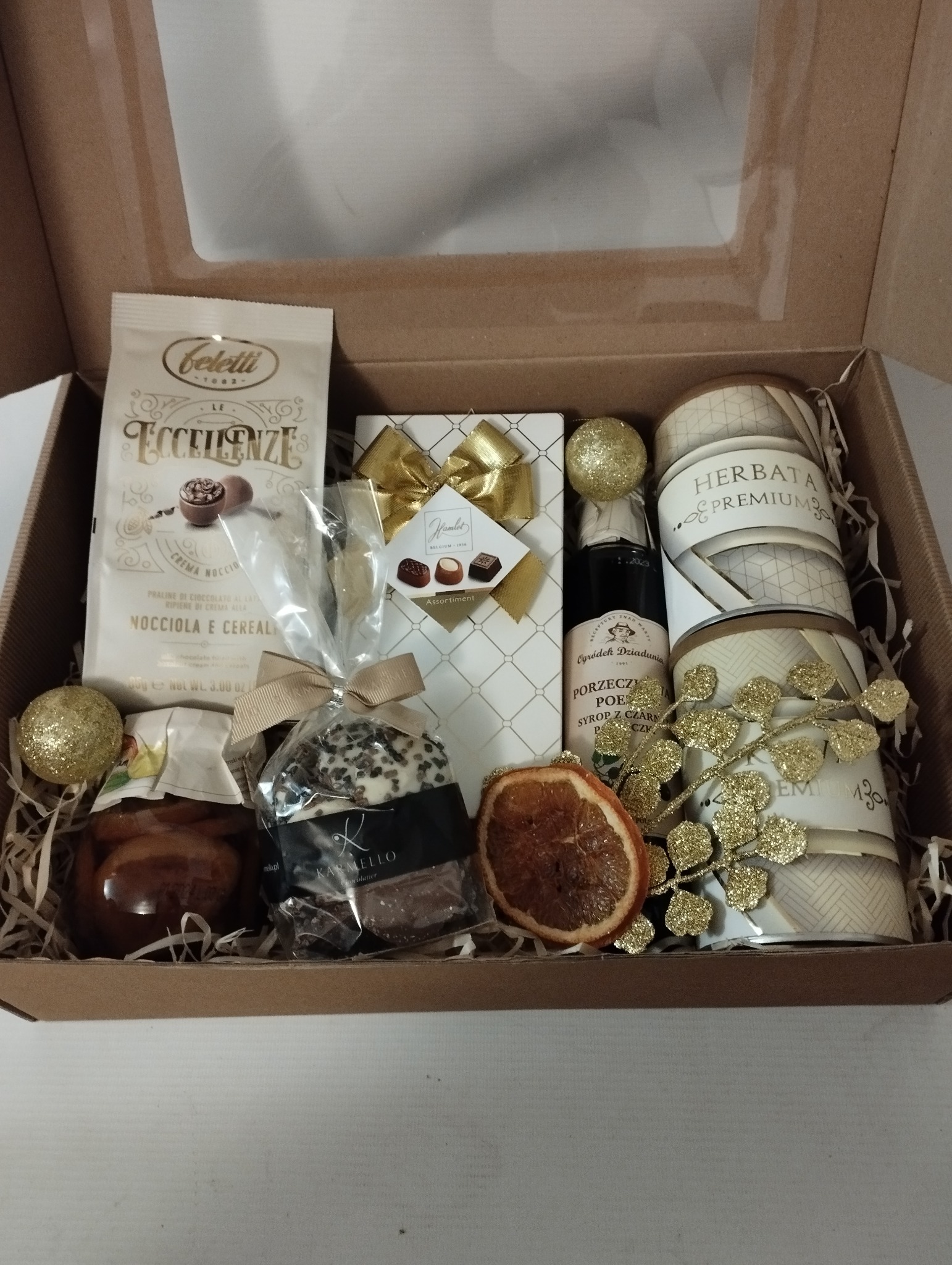 Elegancki kosz prezentowy z kawą, czekoladkami, syropem porzeczkowym, herbatą premium i suszoną pomarańczą, zapakowany w tekturowe pudełko z okienkiem, wypełniony dekoracyjnym siankiem.