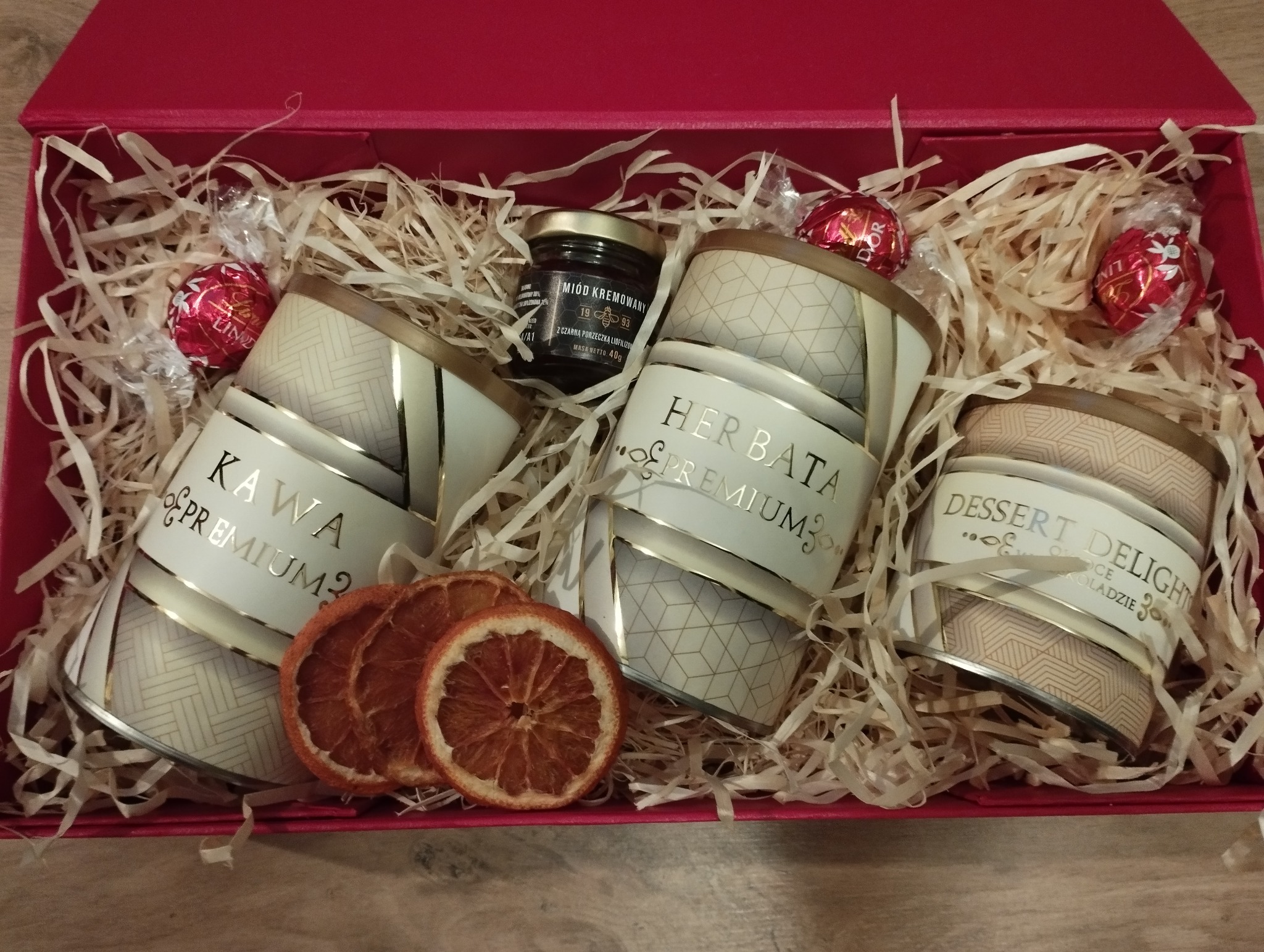 Czerwony kosz prezentowy wypełniony ozdobną wełną drzewną, zawierający trzy puszki z kawą, herbatą i deserem czekoladowym premium, mały słoiczek kremowanego miodu z czarną porzeczką, trzy...