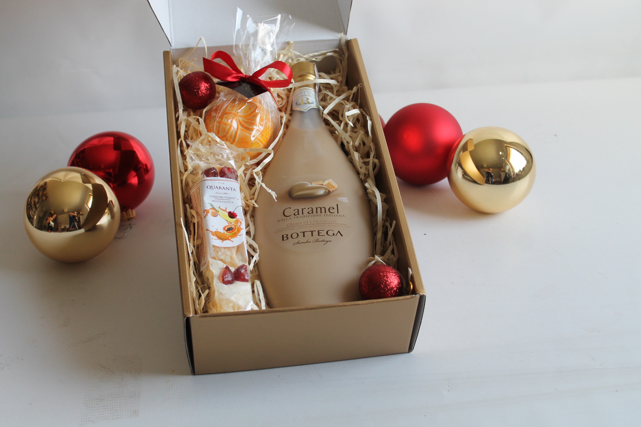 Otwarty kartonowy kosz prezentowy wypełniony butelką likieru karmelowego Bottega, torronem Quarantą, pomarańczą zawiązaną czerwoną wstążką i dekoracyjnymi bombkami świątecznymi na białym tle.