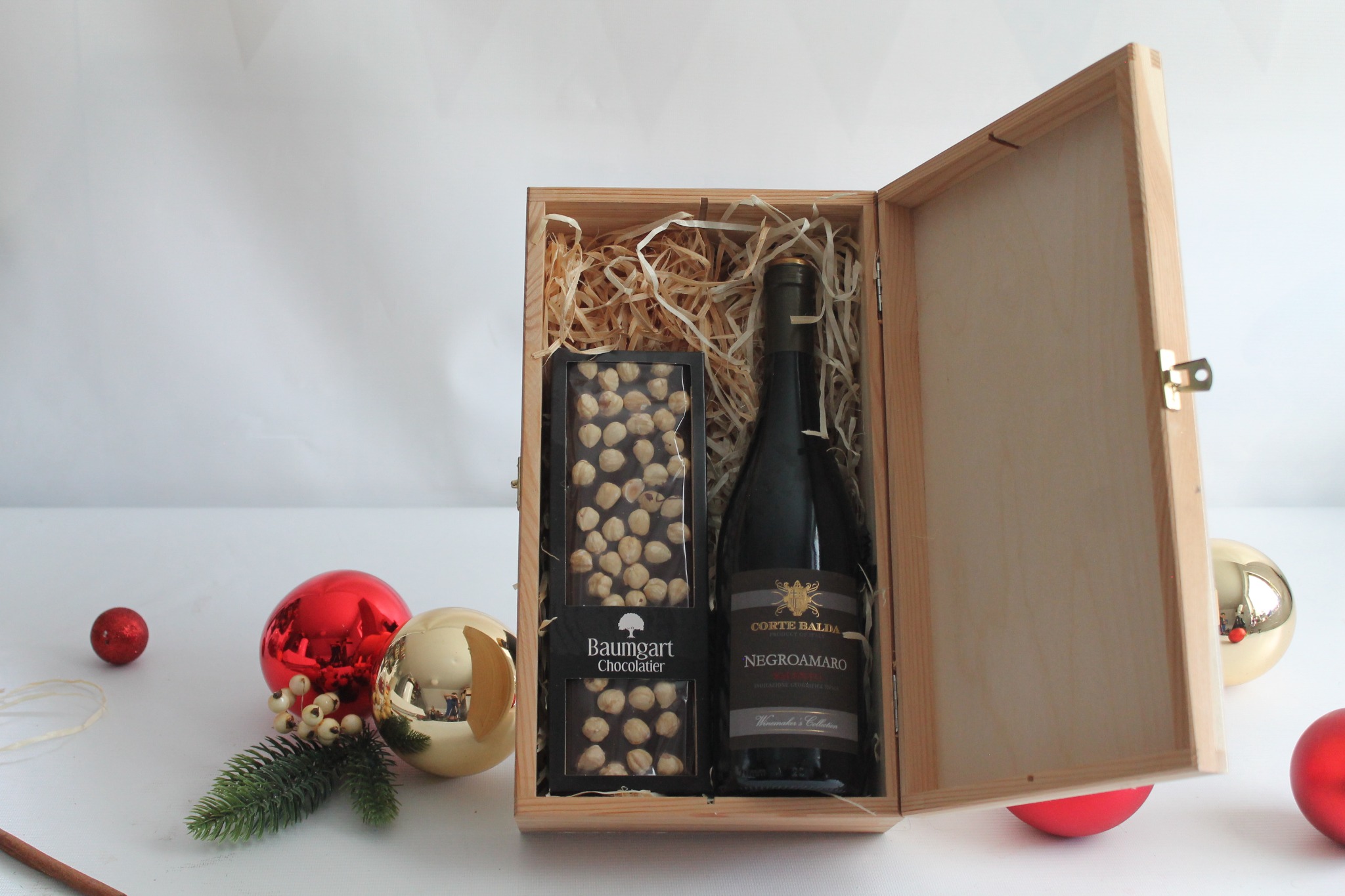 Otwarty drewniany kosz prezentowy wypełniony papierowym wypełniaczem, z butelką wina Negroamaro Corte Balda i czekoladą Baumgart Chocolatier z orzechami laskowymi, ozdobiony bombkami i gałązką...