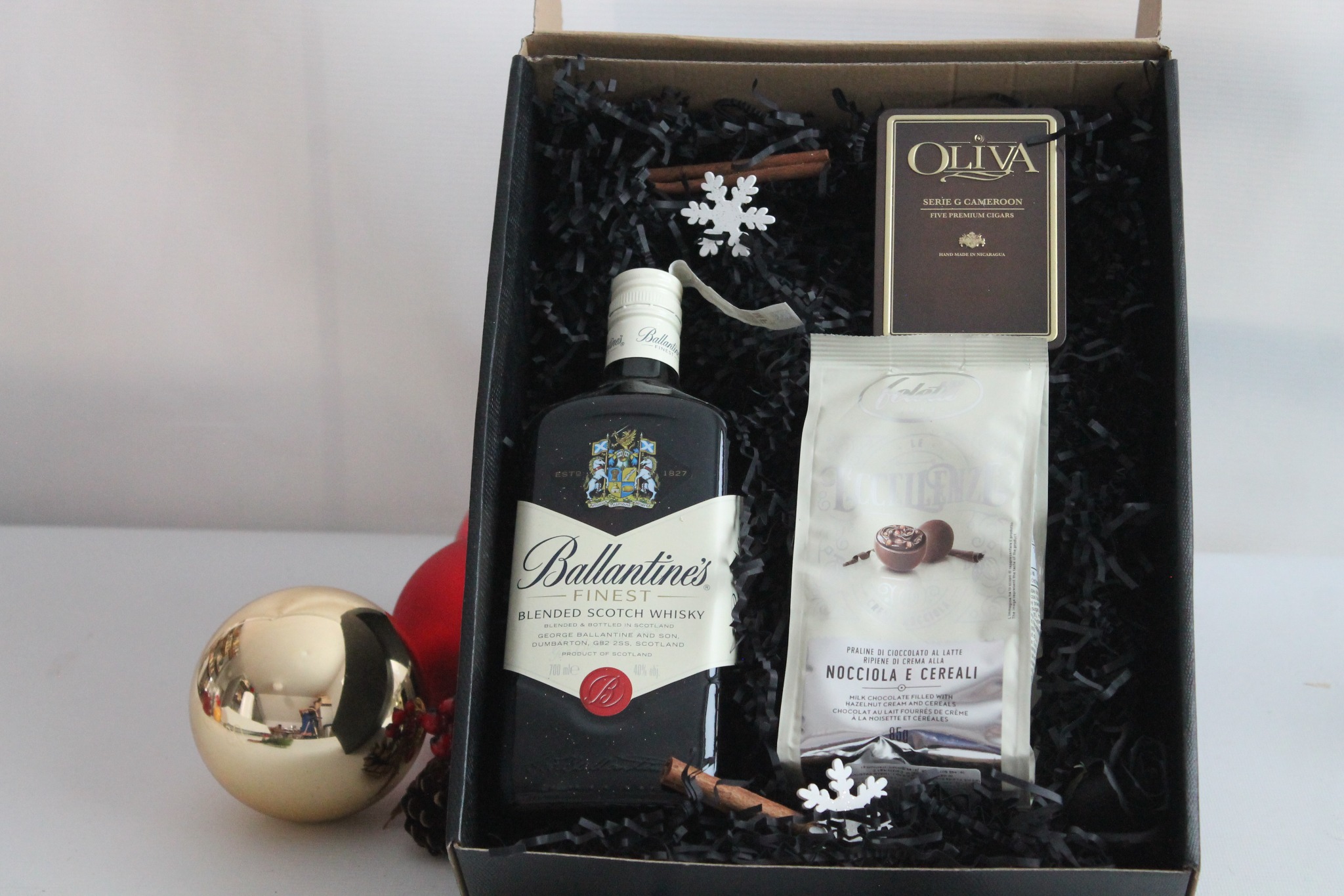 Czarny kosz prezentowy z butelką whisky Ballantine's, czekoladą Oliva, pralinami z orzechami laskowymi i płatkami zbożowymi, ozdobiony cynamonem i sztucznymi śnieżynkami, z bombkami w tle.