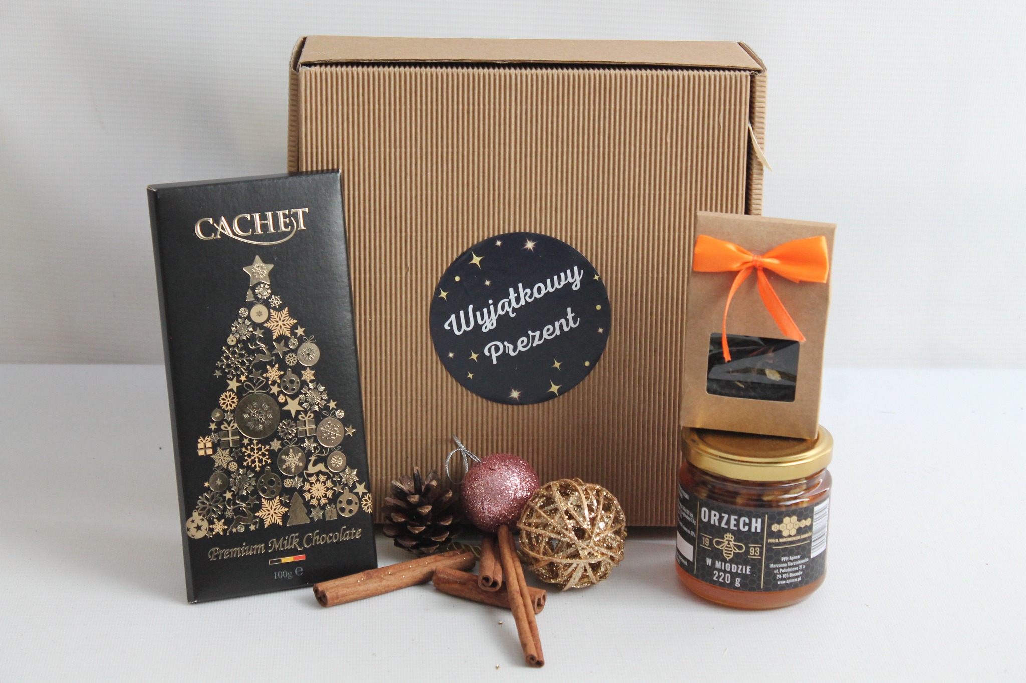 Kartonowe pudełko z naklejką 'Wyjątkowy Prezent' obok czekolady Cachet z motywem choinki, słoika orzechów w miodzie, małej torebki z pomarańczową wstążką, szyszki, dwóch bombek i lasek cynamonu...