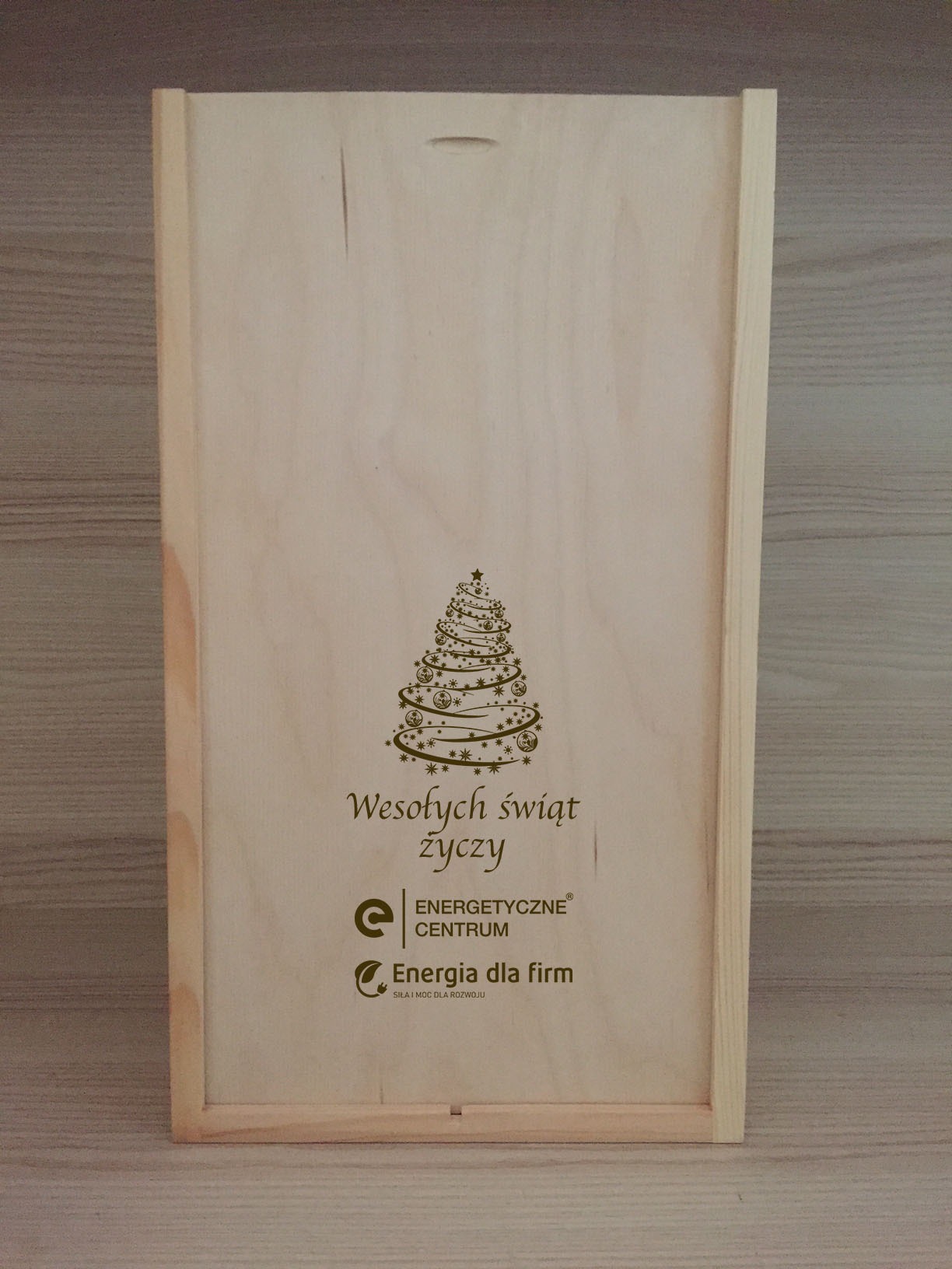 Drewniana skrzynka prezentowa z wygrawerowanym motywem choinki, życzeniami świątecznymi oraz logotypami firmy Energetyczne Centrum i Energia dla firm.