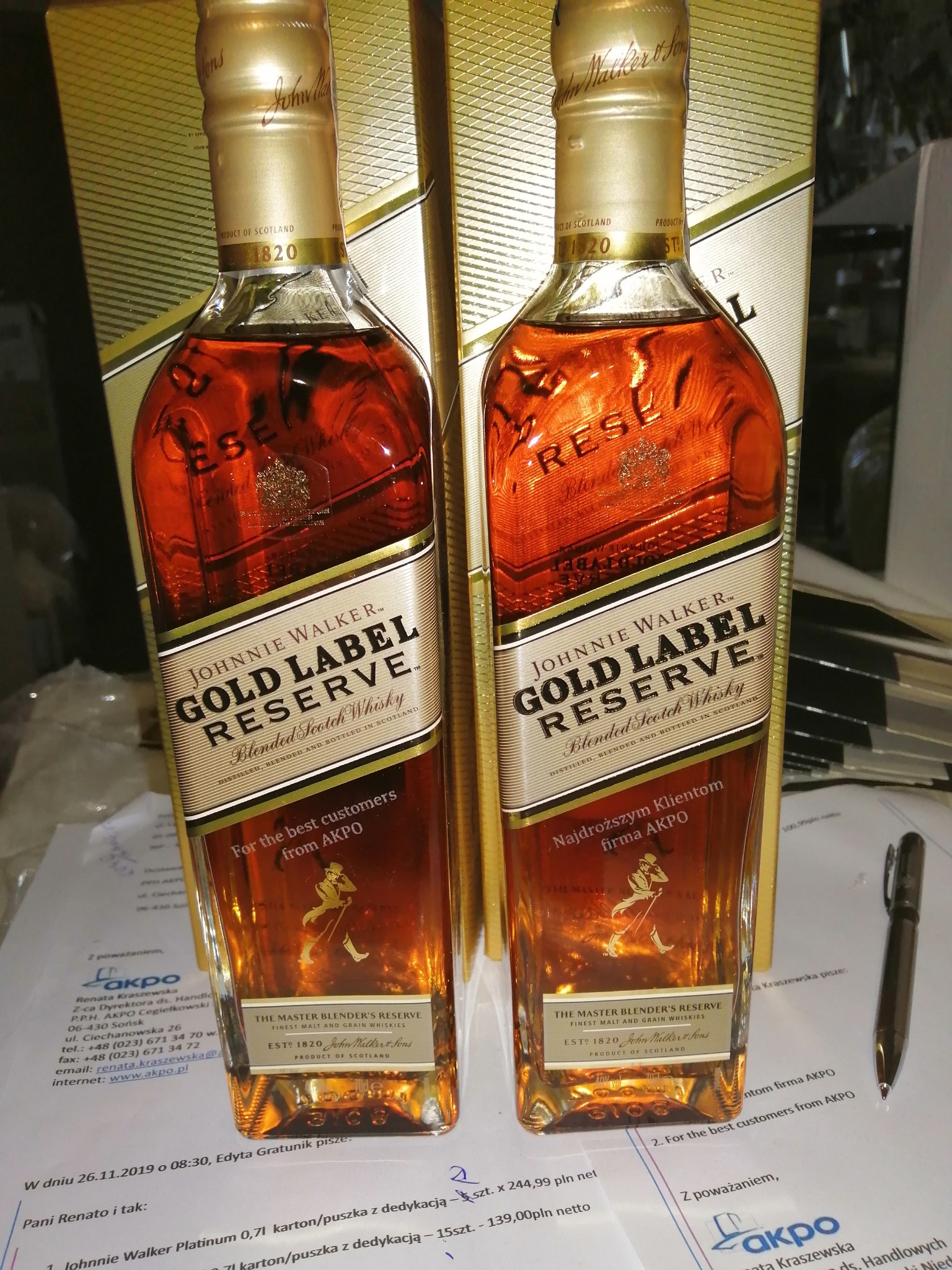 Dwie butelki Johnnie Walker Gold Label Reserve z dedykacją dla firmy AKPO, ustawione na fakturze z zamówieniem, z widocznym złotym opakowaniem w tle i długopisem obok.