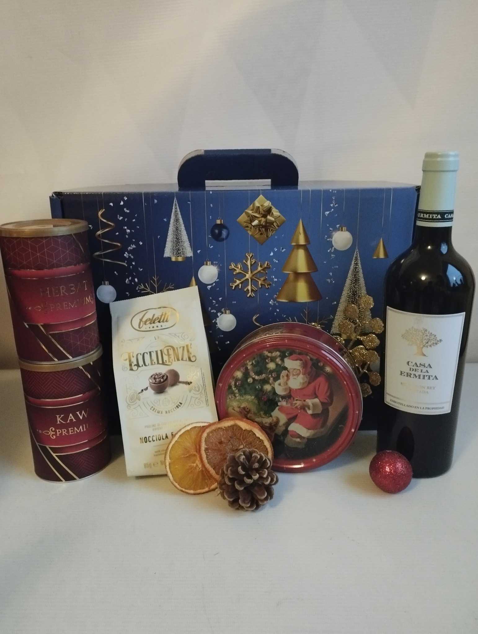 Świąteczny kosz prezentowy z winem, kawą, herbatą, czekoladkami i ozdobami, zapakowany w pudełko z motywem choinek i bombek.