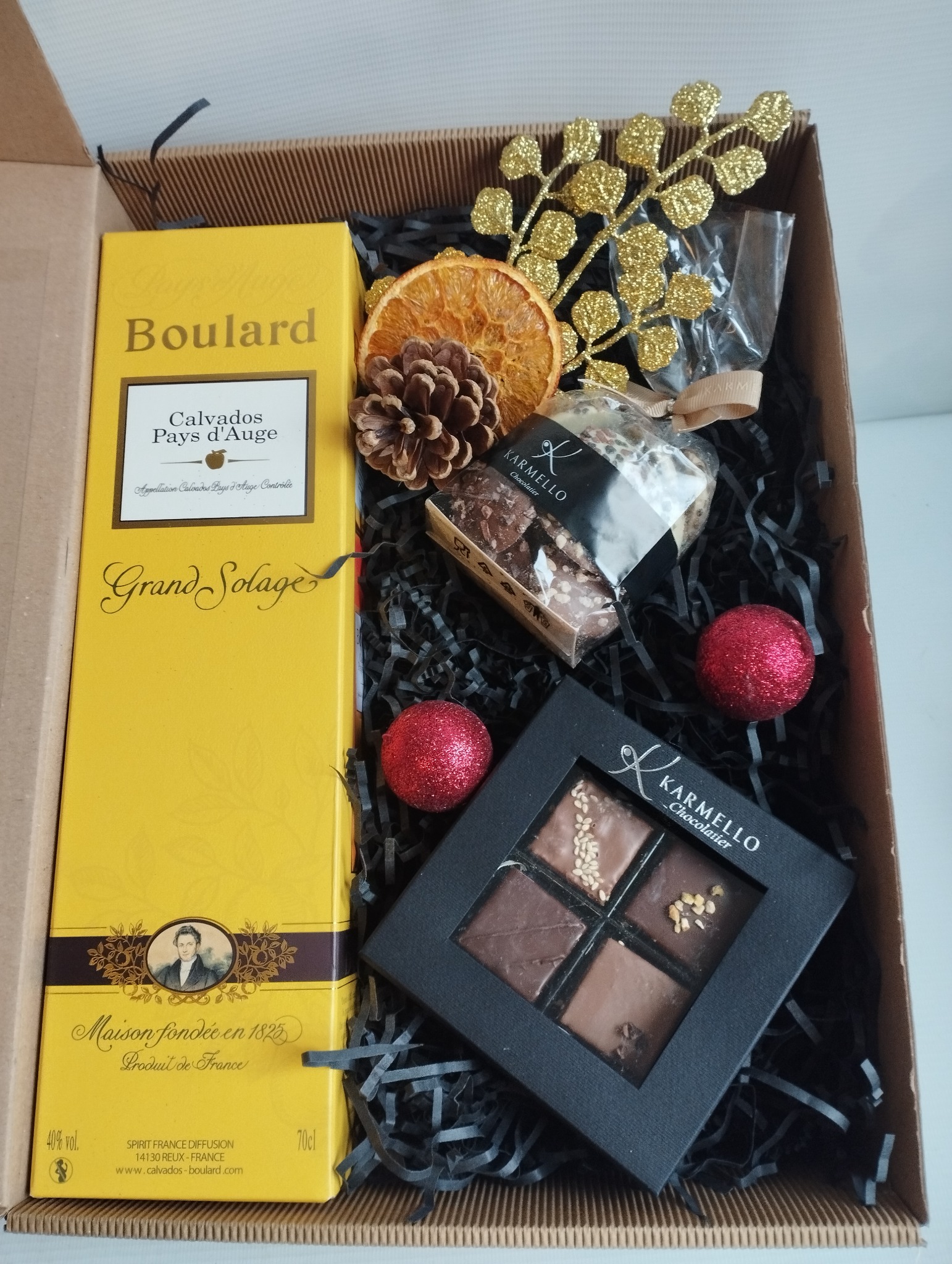 Elegancki kosz prezentowy z Calvadosem Boulard, czekoladkami Karmello, suszoną pomarańczą, szyszką i złotą ozdobą na czarnym wypełniaczu w kartonowym pudełku.