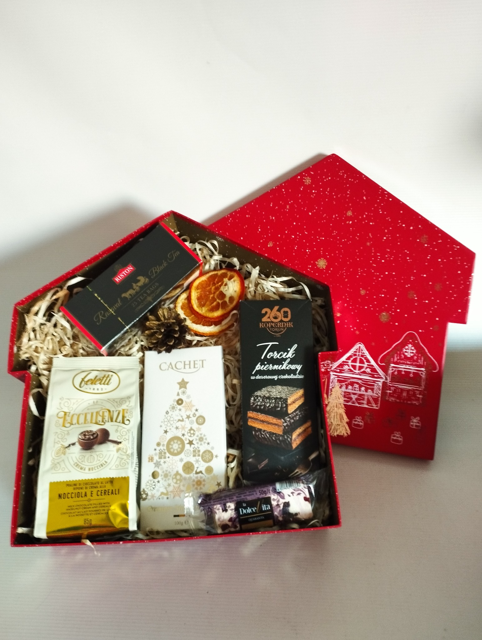 Czerwony, świąteczny kosz prezentowy w kształcie domku, wypełniony kawą, herbatą, piernikami, czekoladą i suszonymi pomarańczami, ułożony na białym tle.