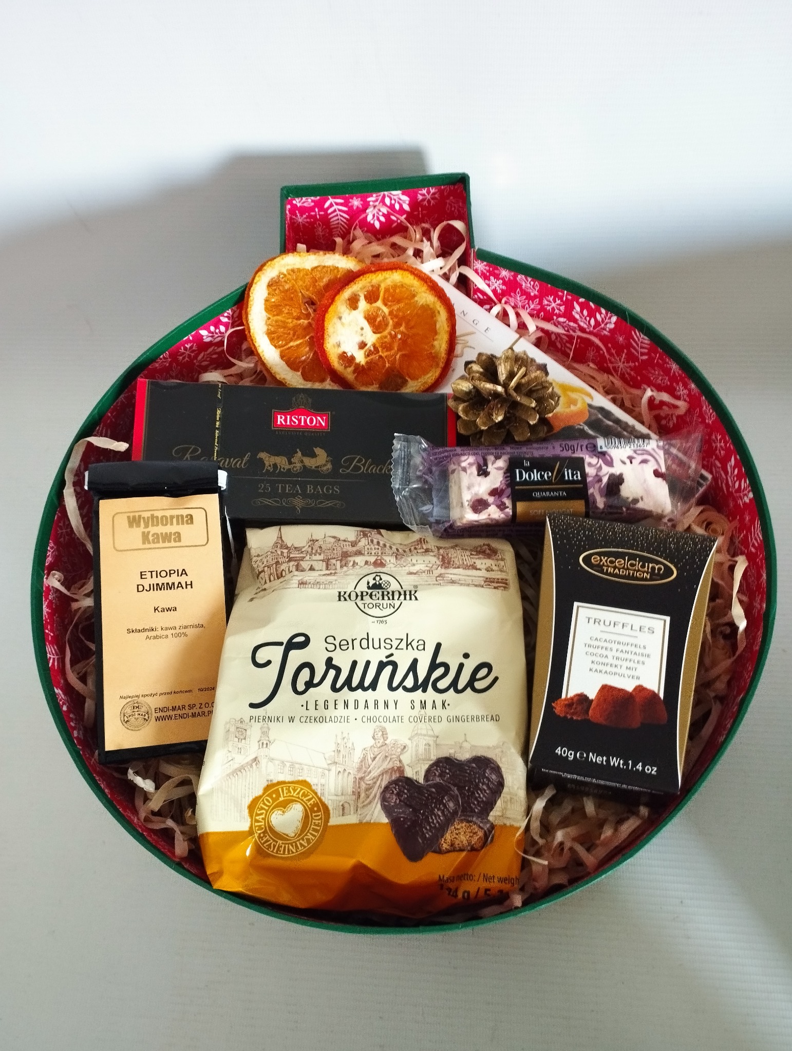 Okrągły, zielono-czerwony kosz z zawartością: kawa Etiopia Djimmah, czarna herbata Riston, pierniki w czekoladzie Serduszka Toruńskie, trufle Excelcium Tradition, nugat Dolce Vita Quaranta, dwie...
