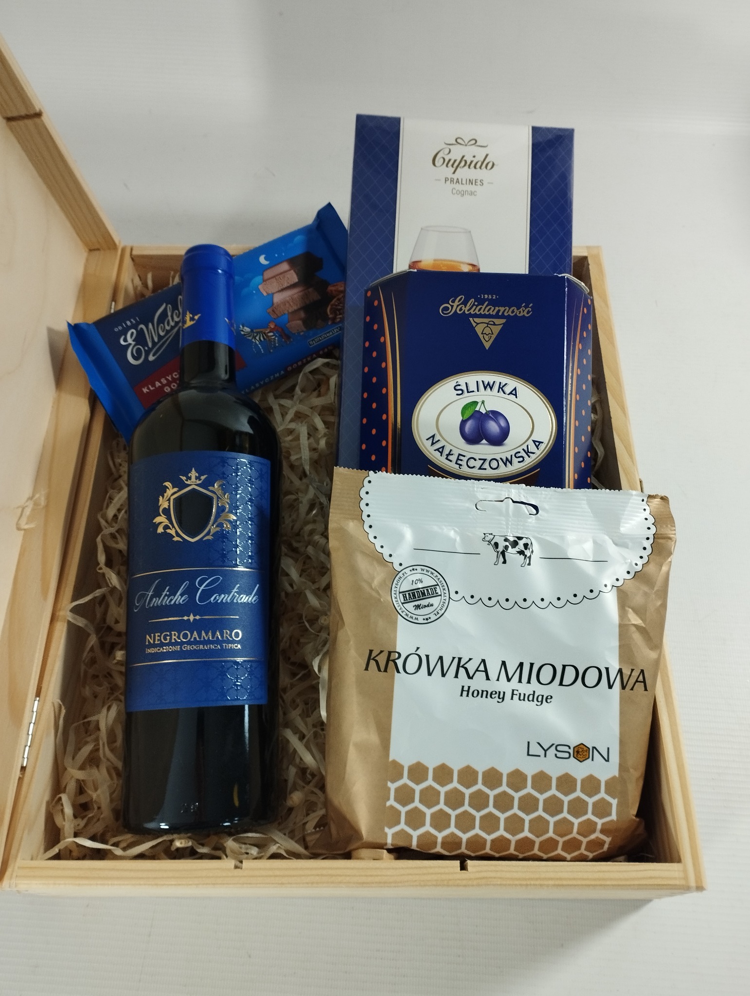 Drewniany kosz wypełniony słodyczami, butelką wina Negroamaro Antiche Contrade, pralinkami Cupido Cognac, śliwkami w czekoladzie Solidarność i krówkami miodowymi Lysson, całość wyłożona...