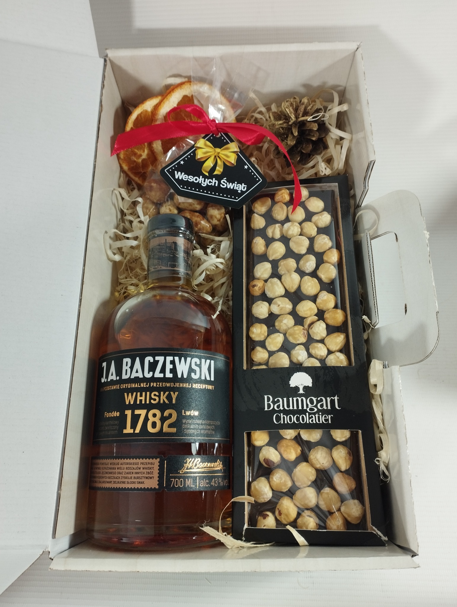 Elegancki kosz prezentowy z butelką whisky J.A. Baczewski, czekoladą Baumgart z orzechami laskowymi, kandyzowanymi pomarańczami i dekoracyjną szyszką, wypełniony papierowym siankiem i ozdobiony...