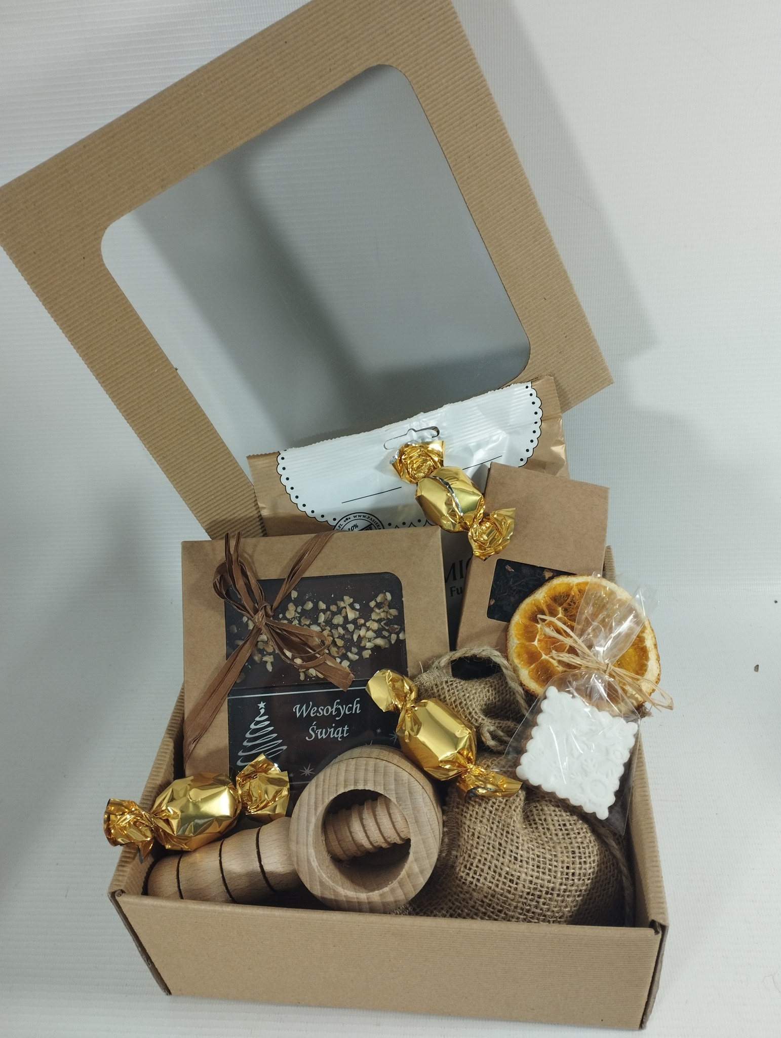 Otwarty kosz prezentowy z kartonu z okienkiem, wypełniony słodyczami w złotych papierkach, czekoladą z napisem 'Wesołych Świąt', suszoną pomarańczą i drewnianym dziadkiem do orzechów.