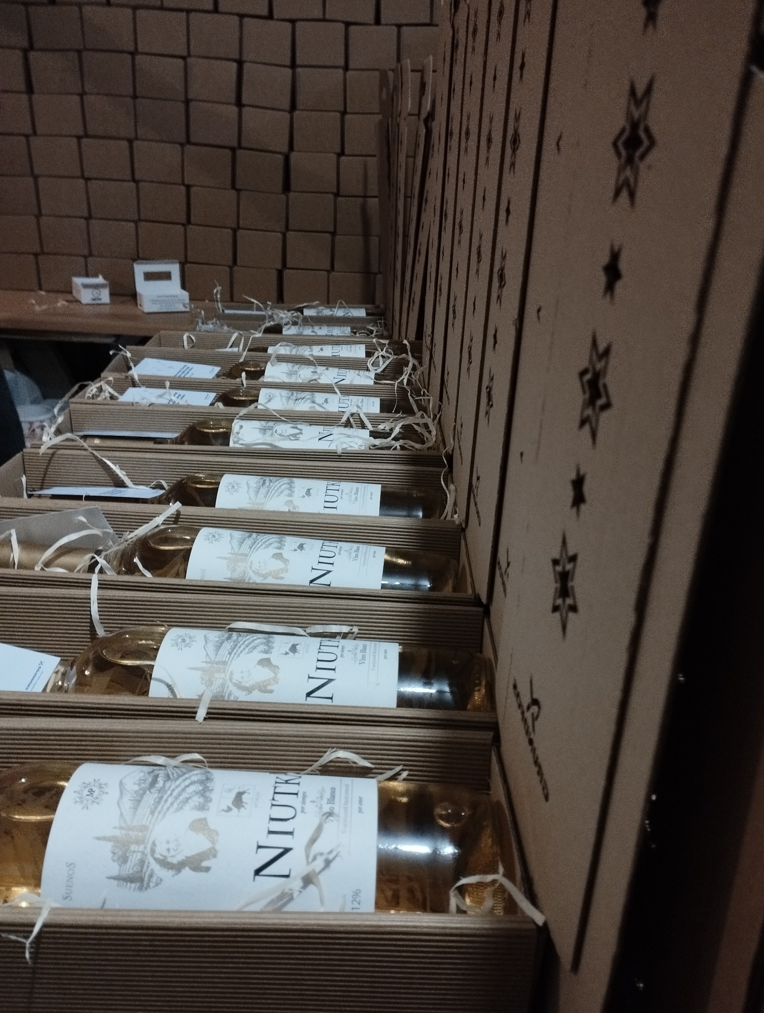 Rząd kartonowych pudełek z butelkami białego wina Niutka, wypełnionych ozdobnym siankiem, ustawionych na tle ściany z kartonów, gotowych do wysyłki.