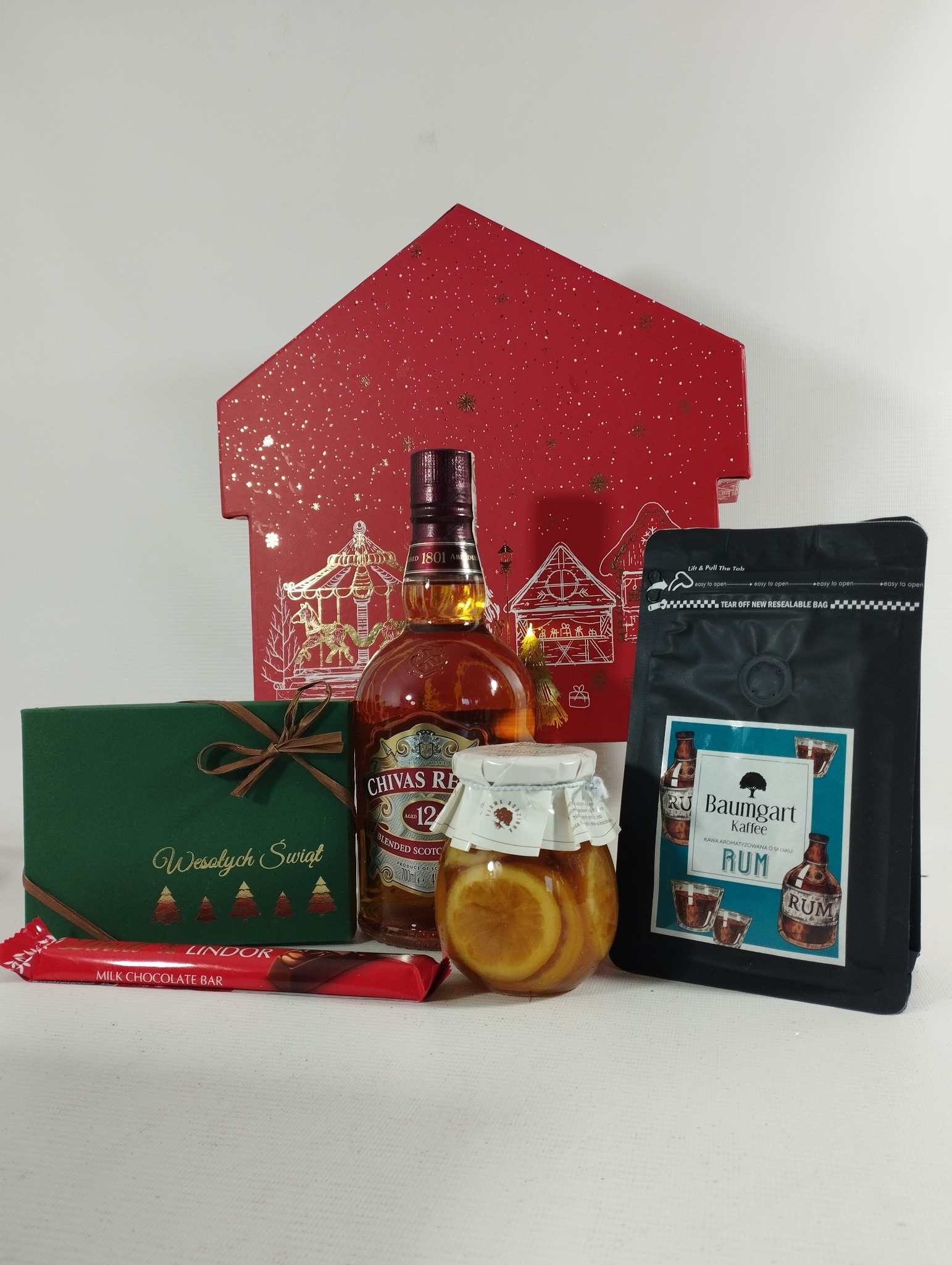 Świąteczny zestaw prezentowy: butelka whisky Chivas Regal 12, słoik z cytrynami w syropie, kawa aromatyzowana o smaku rumu Baumgart Kaffe, czekolada Lindor i zielone pudełko z napisem 'Wesołych...