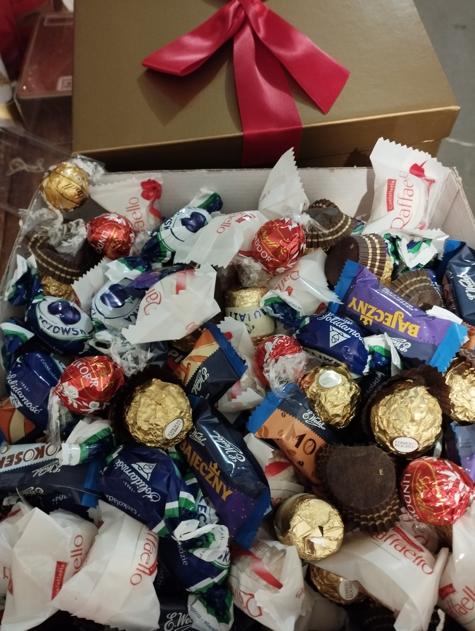 Otwarty kartonowy kosz prezentowy wypełniony różnorodnymi słodyczami w opakowaniach, w tym czekoladki Raffaello, Ferrero Rocher i Wedel, z czerwoną wstążką na górze.
