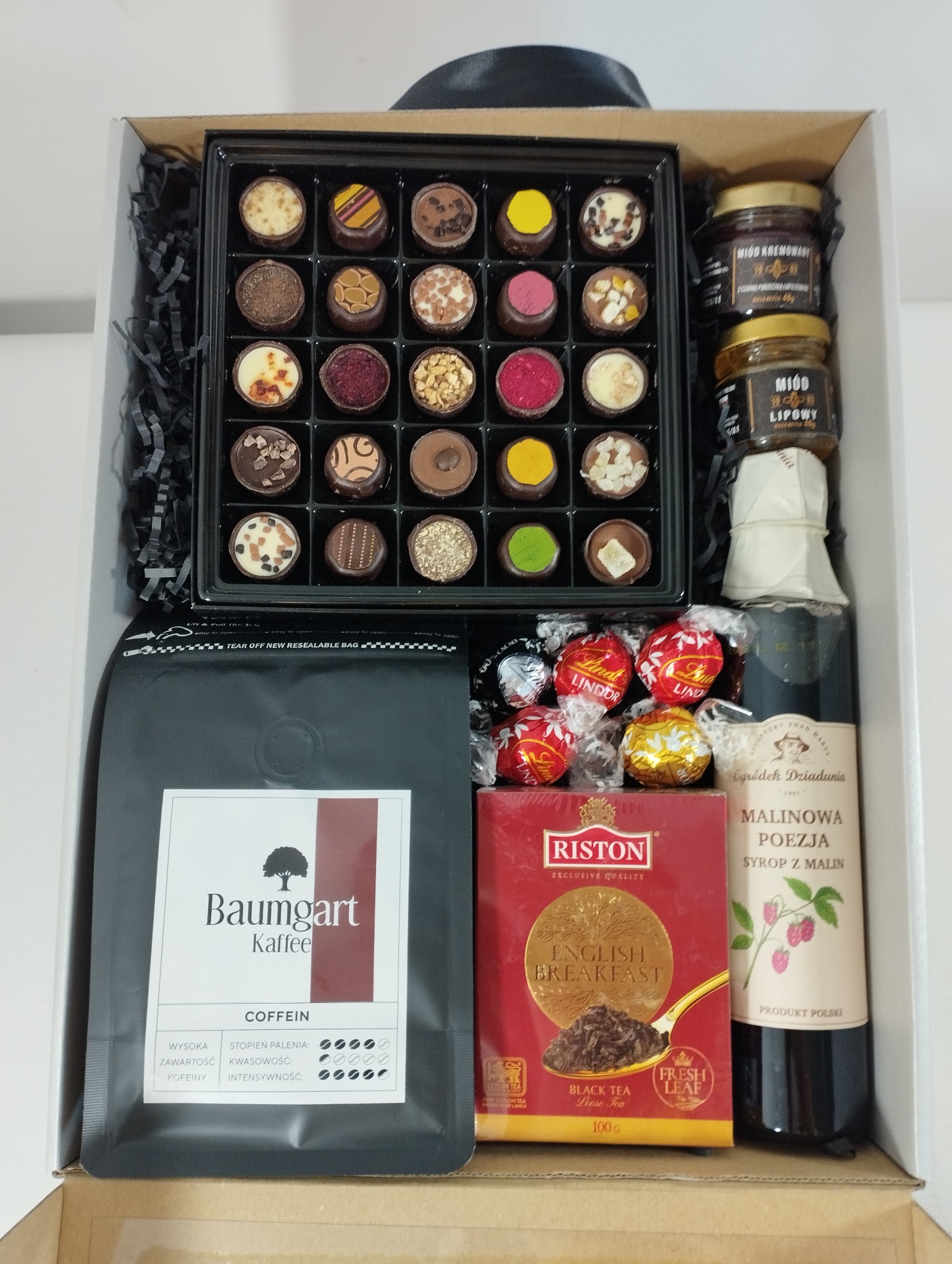 Otwarty biały karton z czarną wstążką wypełniony koszem prezentowym: czekoladki w czarnym pudełku, miody w słoiczkach, kawa Baumgart, herbata Riston, syrop malinowy Ogródek Dziadunia i czekoladki...