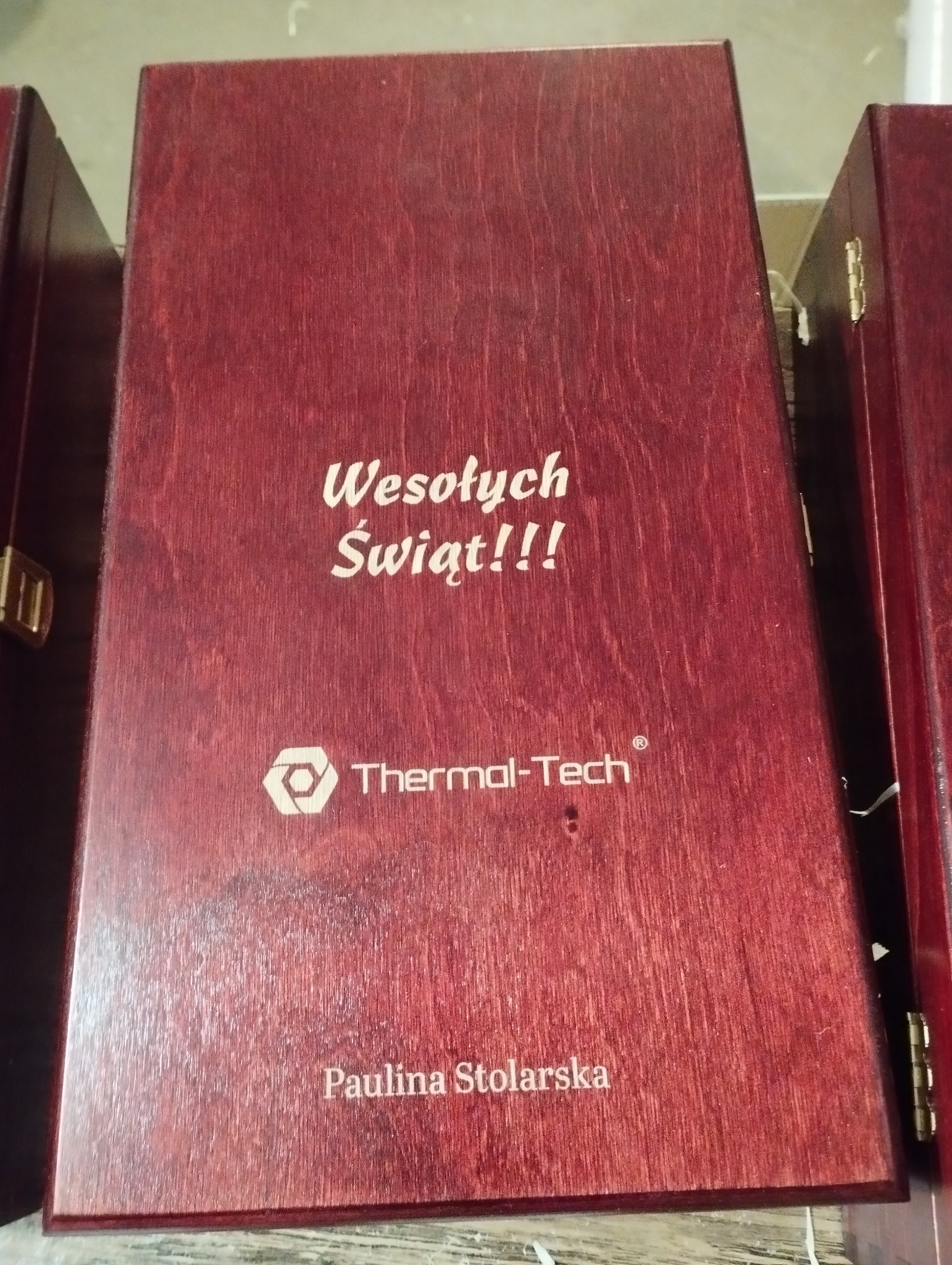Ciemnoczerwone, drewniane pudełko z napisem 'Wesołych Świąt!!!', logo 'Thermal-Tech' i imieniem 'Paulina Stolarska', stojące obok podobnych pudełek.