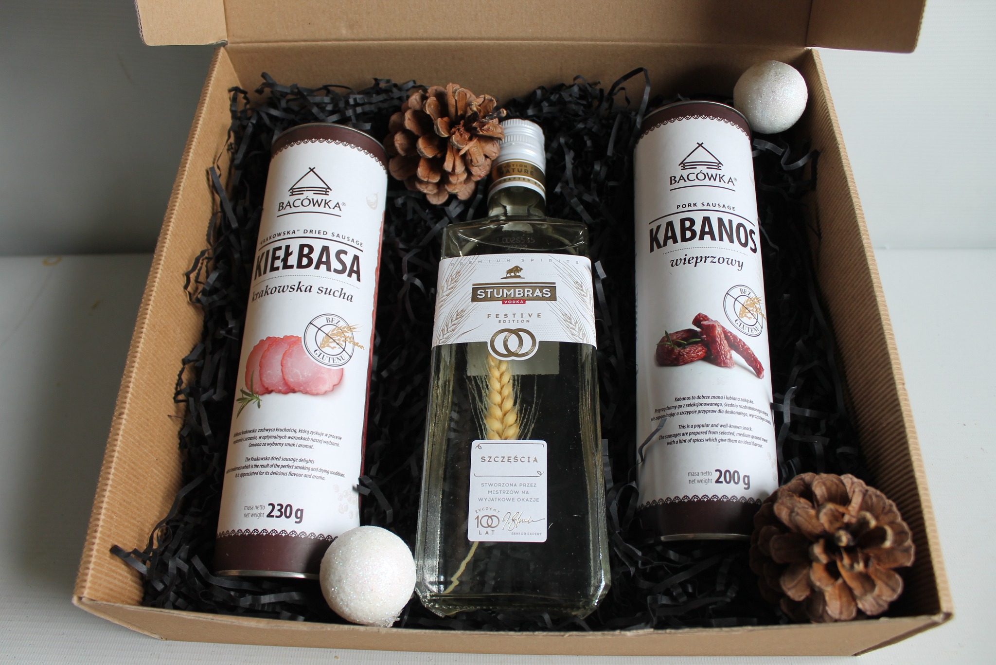 Elegancki kosz prezentowy: kabanosy, kiełbasa krakowska, wódka Stumbras Festive Edition, szyszki i błyszczące kule w kartonowym pudełku z czarnym wypełniaczem.