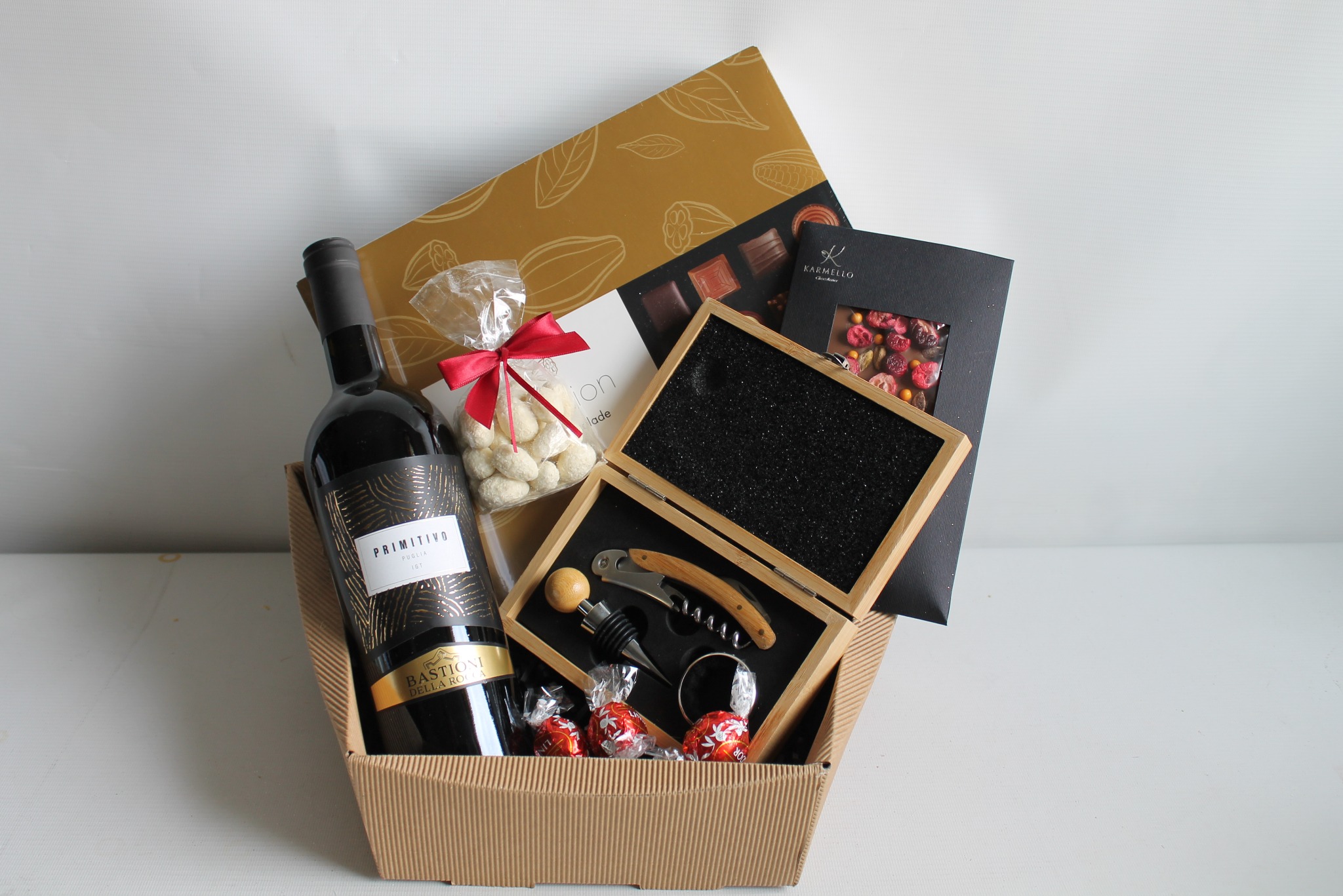 Elegancki kosz prezentowy z winem Primitivo, czekoladkami Karmello, orzechami w lukrze i akcesoriami do wina w drewnianym pudełku, umieszczony w tekturowym koszu.