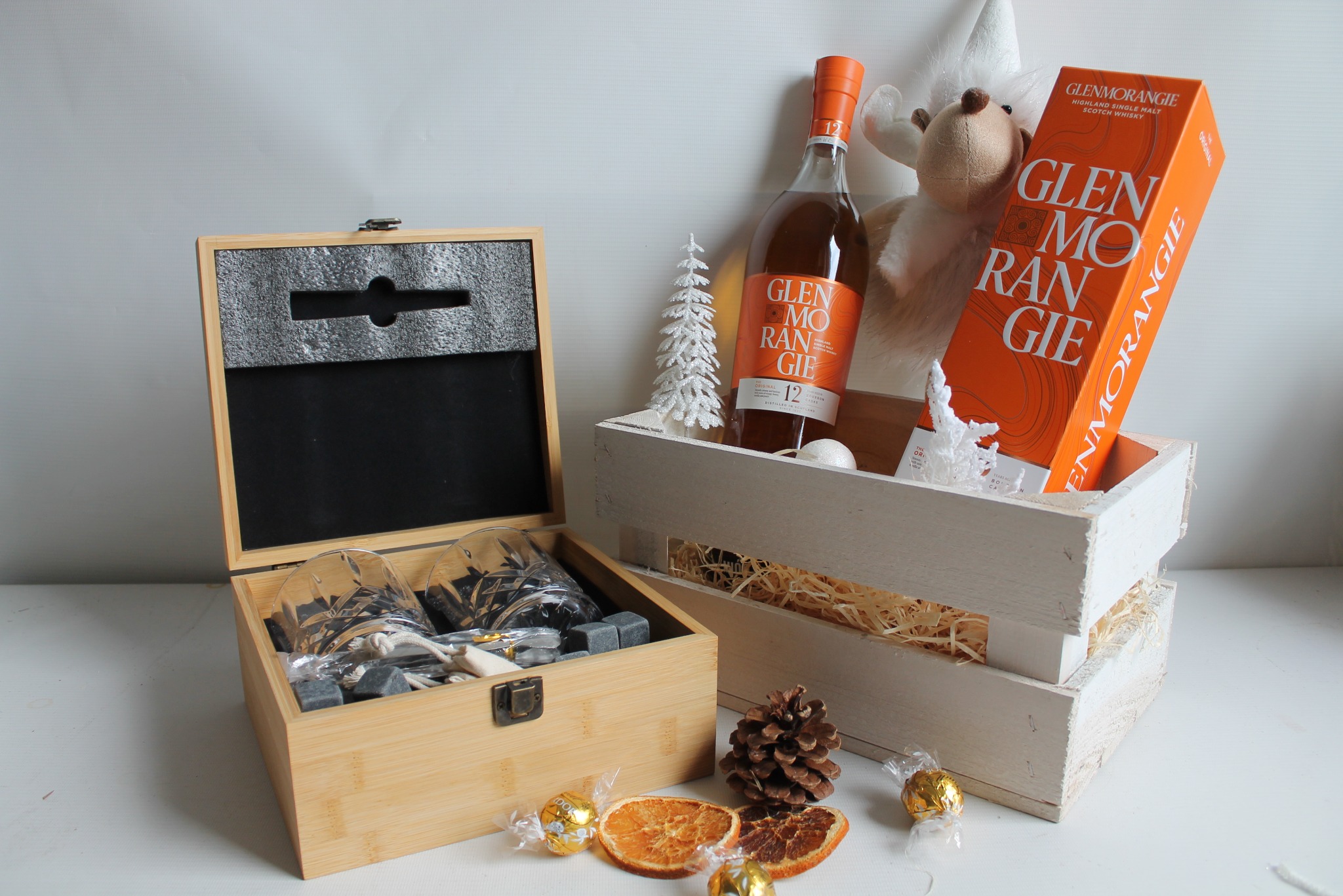 Elegancki kosz prezentowy z whisky Glenmorangie, szklankami i kamieniami do chłodzenia w drewnianych skrzynkach, ozdobiony suszonymi pomarańczami i słodyczami.