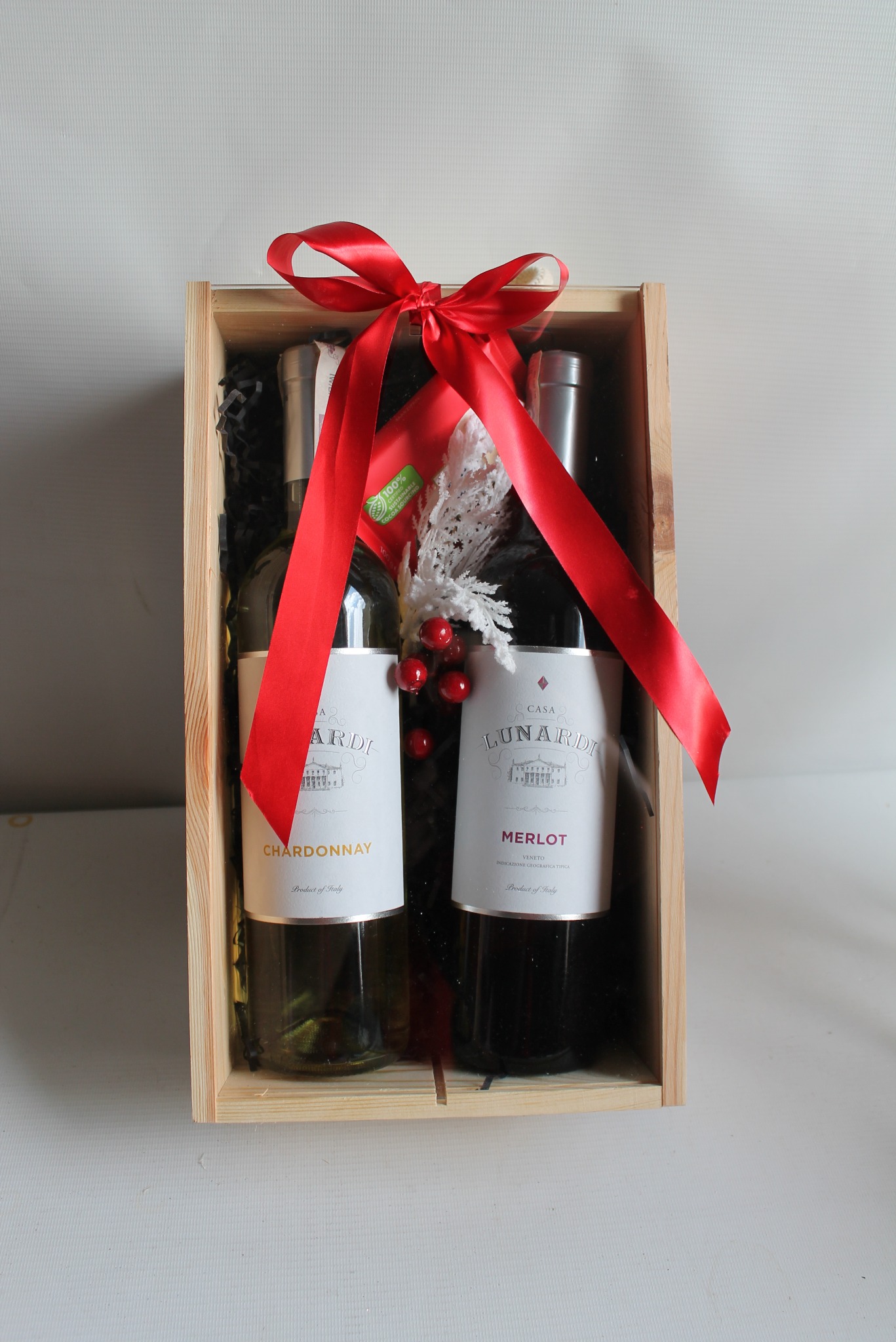 Elegancki kosz prezentowy: drewniana skrzynka z winem Chardonnay i Merlot, ozdobiona czerwoną wstążką i dekoracją z białych gałązek i czerwonych koralików.