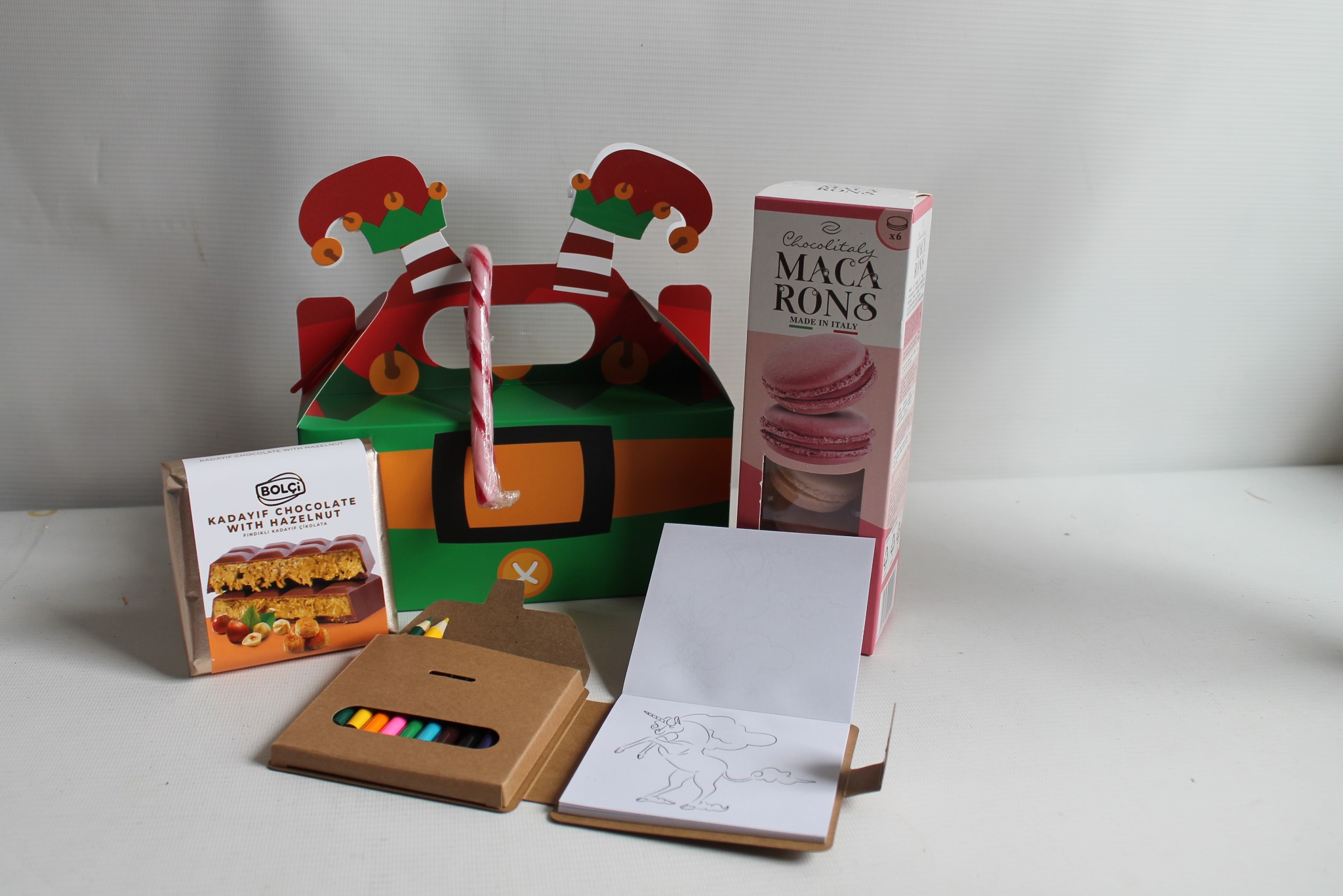 Świąteczny kosz prezentowy z czekoladą, makaronikami i kolorowanką z kredkami, zapakowany w pudełko w kształcie elfa.