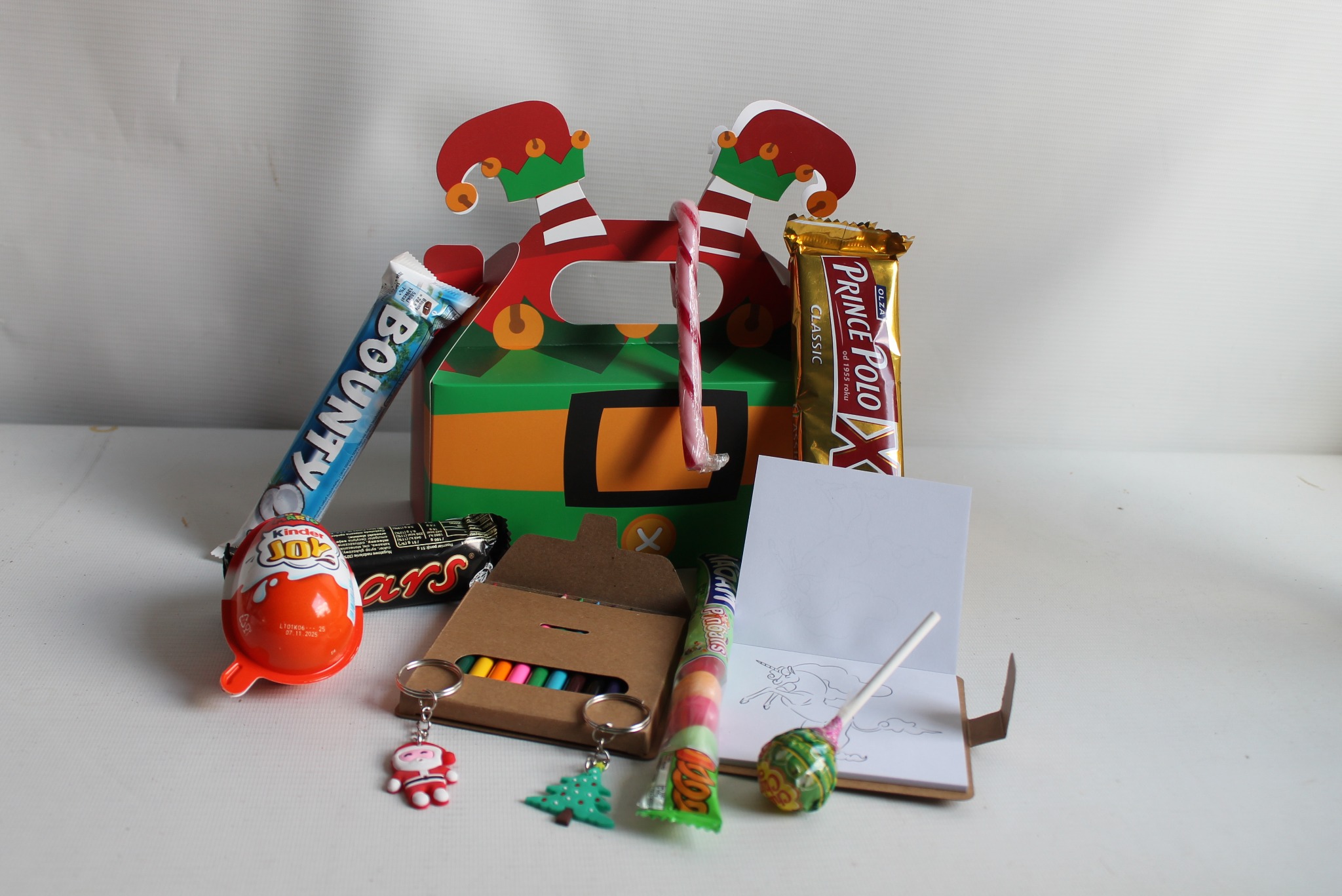 Świąteczny kosz prezentowy z elfim motywem: słodycze, kredki, notatnik z jednorożcem i breloczki na białym tle. Idealny upominek dla dziecka.