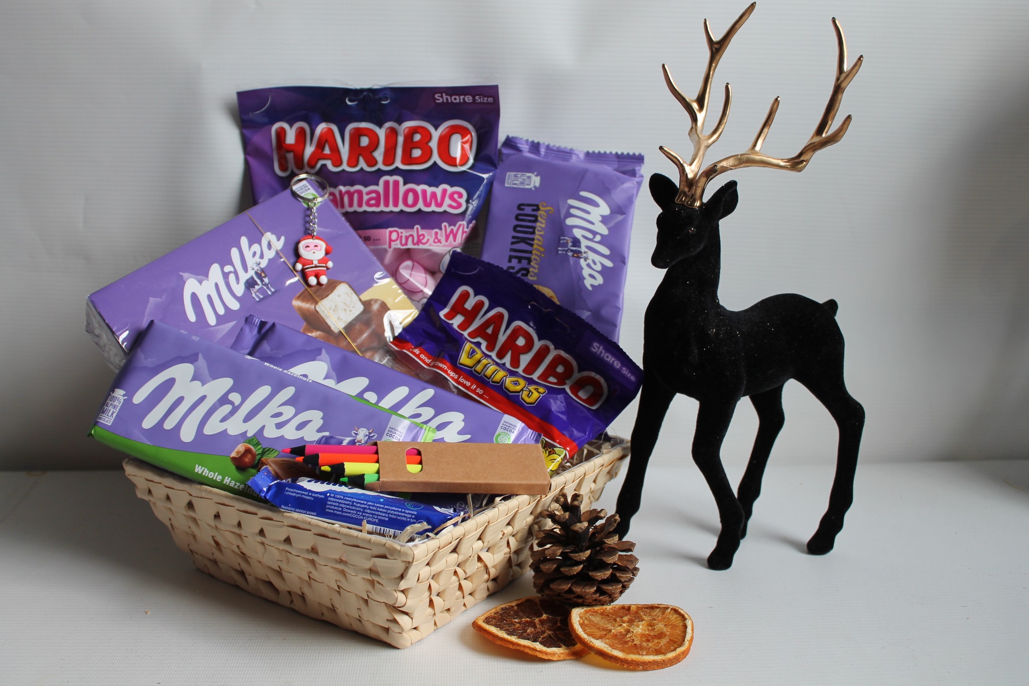 Świąteczny kosz upominkowy: słodycze Milka i Haribo w wiklinowym koszu, obok czarny renifer z brokatem i suszone pomarańcze. Idealny prezent na Boże Narodzenie.