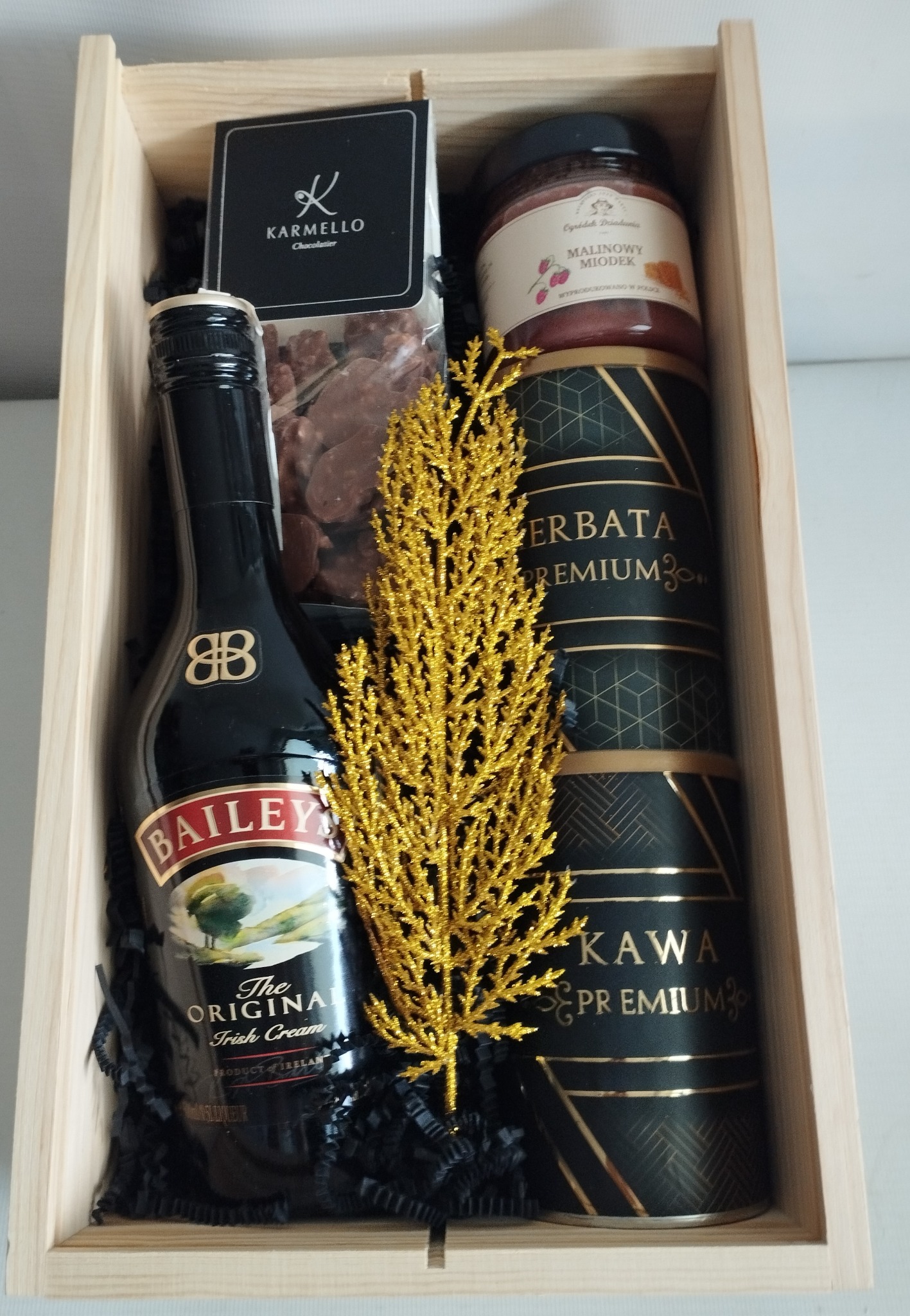 Elegancki kosz prezentowy w jasnym, drewnianym pudełku. Zawiera likier Baileys, miód malinowy, kawę premium, herbatę i czekoladki Karmello, ozdobiony złotą gałązką.