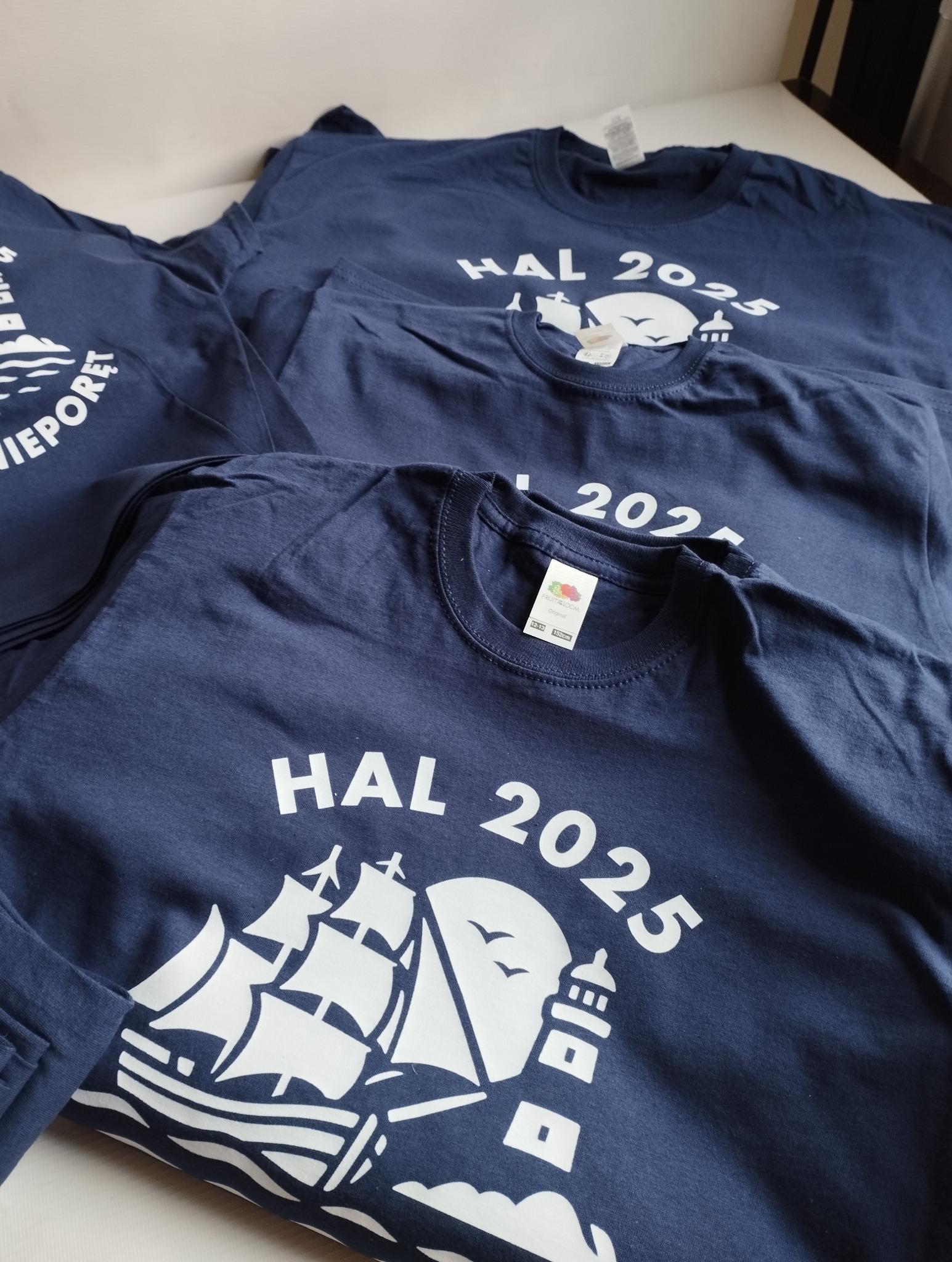 Granatowe koszulki z białym nadrukiem: żaglowiec, latarnia morska i napis HAL 2025. Ułożone jedna na drugiej, widoczne metki. Detaliczny widok na gotowe produkty.