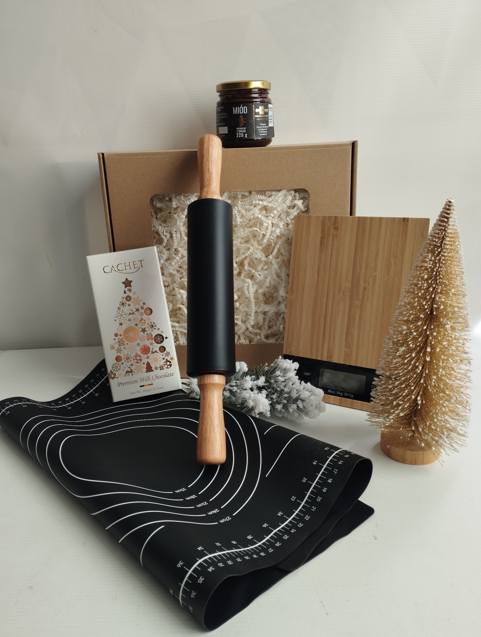 Świąteczny kosz prezentowy: miód z kakao, czekolada Cachet, wałek, mata do ciasta, waga kuchenna i dekoracyjna choinka na białym tle.